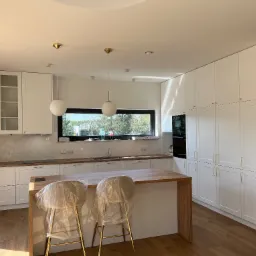 Jasna, nowoczesna kuchnia z białymi szafkami, drewnianym blatem i wyspą; krzesła barowe zabezpieczone folią ochronną.