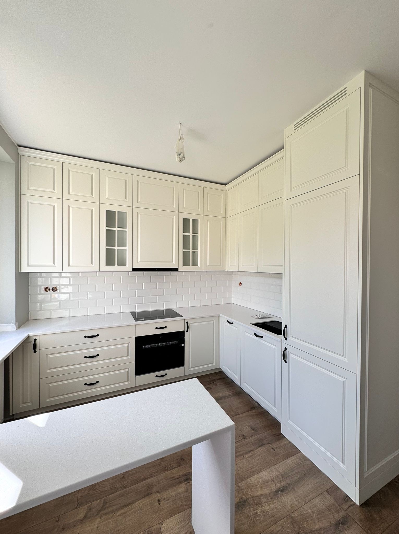 Jasna kuchnia w stylu klasycznym z białymi frontami szafek z frezowaniami, czarnymi uchwytami i blatem w kolorze złamanej bieli. Na ścianie białe płytki typu metro. Widoczne gniazdka elektryczne...