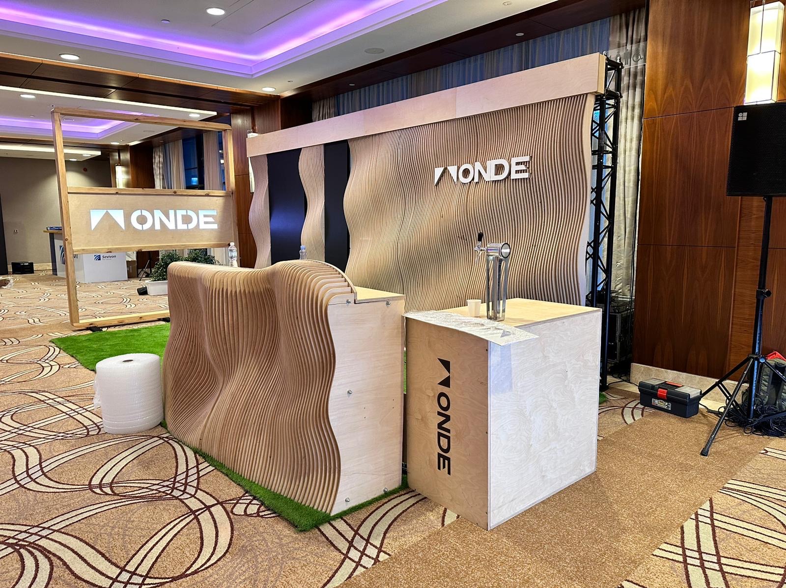 Drewniane stoisko targowe z falistymi elementami i logotypem firmy, uzupełnione o drewniany bar z nalewakiem do piwa, umieszczone na wykładzinie w sali konferencyjnej.