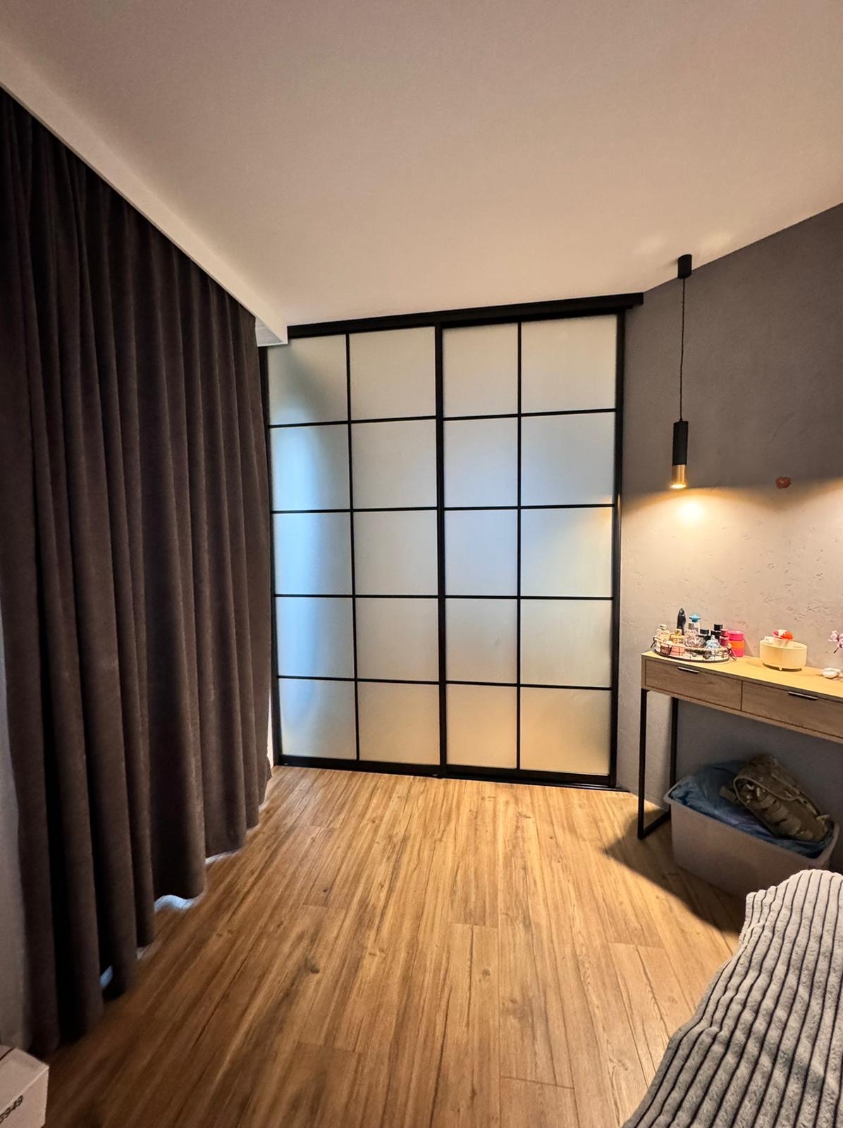 Nowoczesna szafa przesuwna z matowego szkła i czarnymi szprosami, wkomponowana w aranżację sypialni z drewnianą podłogą i toaletką pod wiszącą lampą.