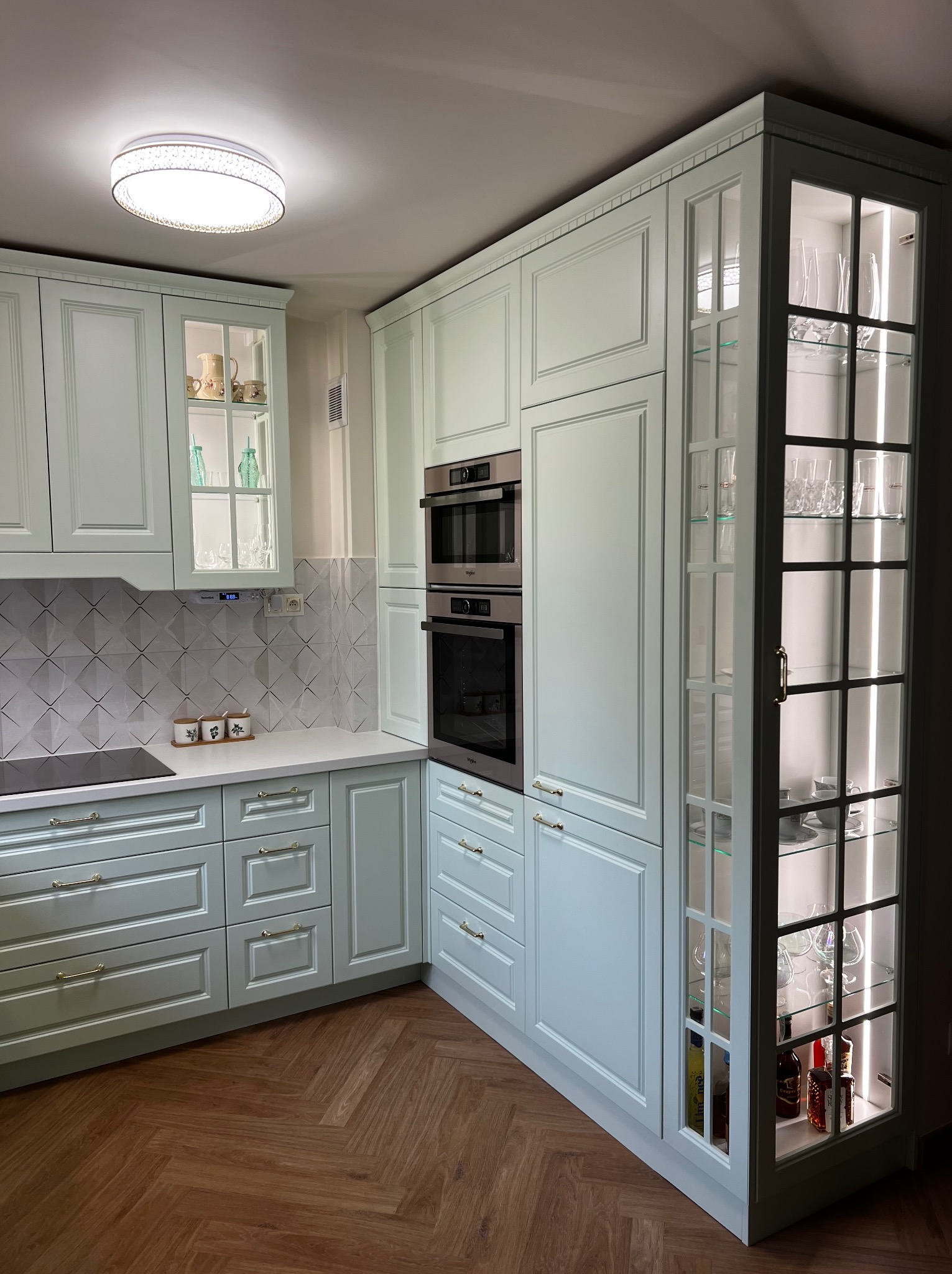 Jasna, pastelowa kuchnia na wymiar z drewnianą podłogą w jodełkę, przeszklone szafki z podświetleniem LED eksponujące szkło i butelki, zabudowane piekarniki.