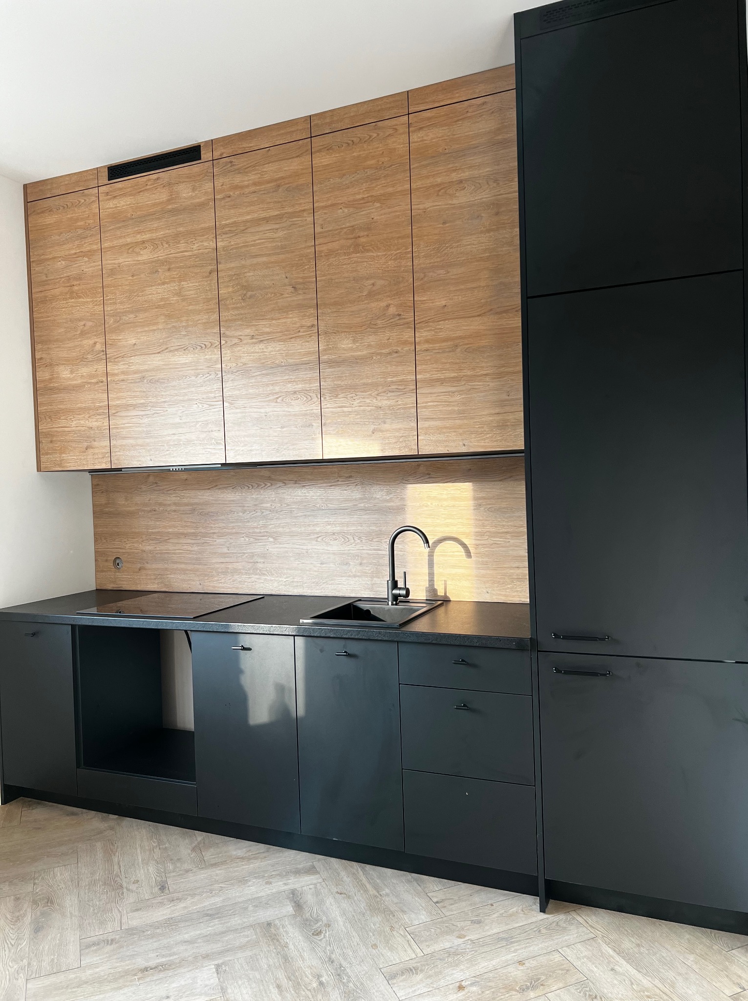 Nowoczesna kuchnia z czarnymi dolnymi szafkami i drewnopodobnymi górnymi szafkami, czarny blat i armatura, parkiet w jodełkę na podłodze.