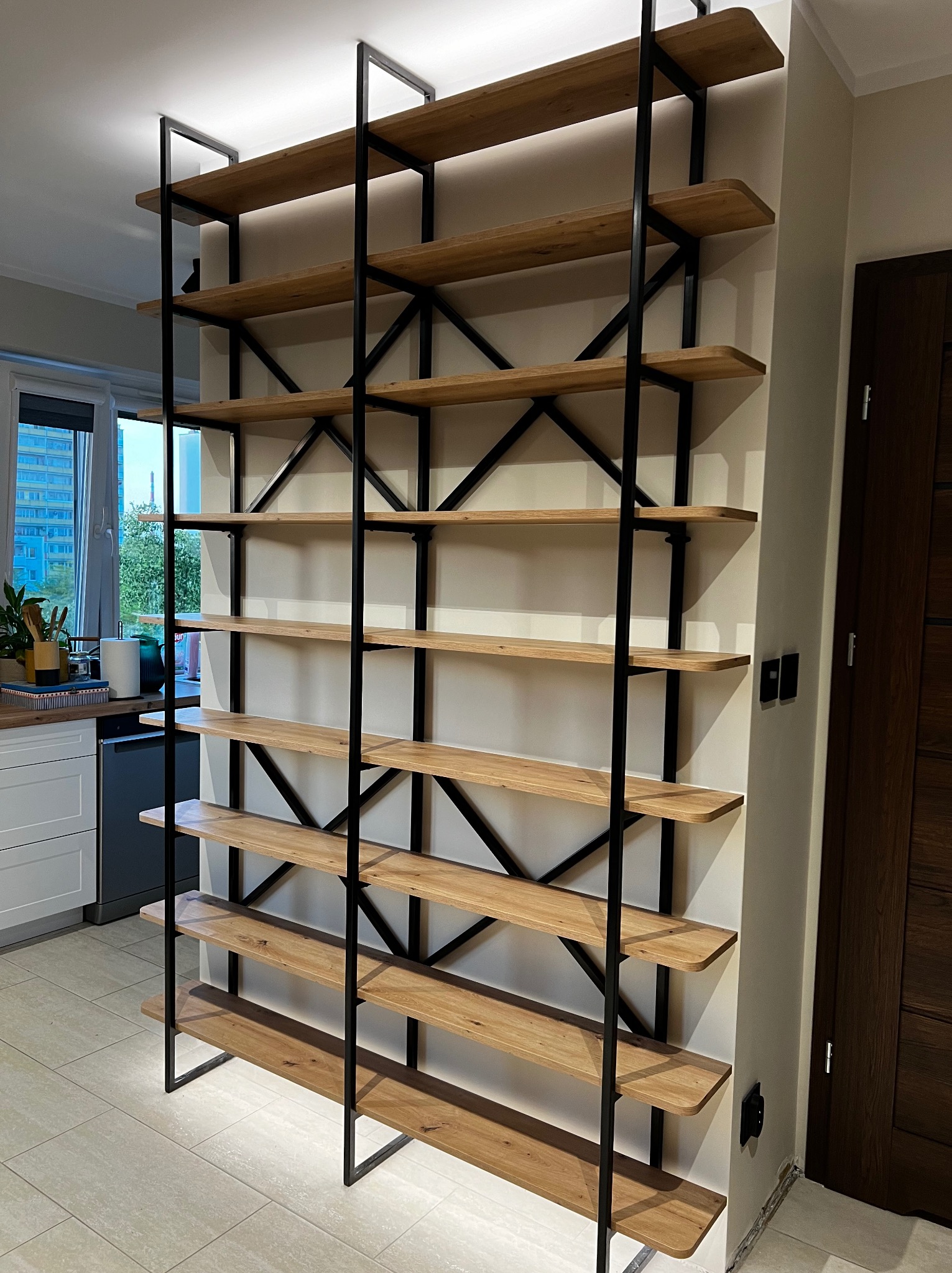 Regał z drewnianymi półkami i czarną metalową konstrukcją, w tle widoczna kuchnia i okno.