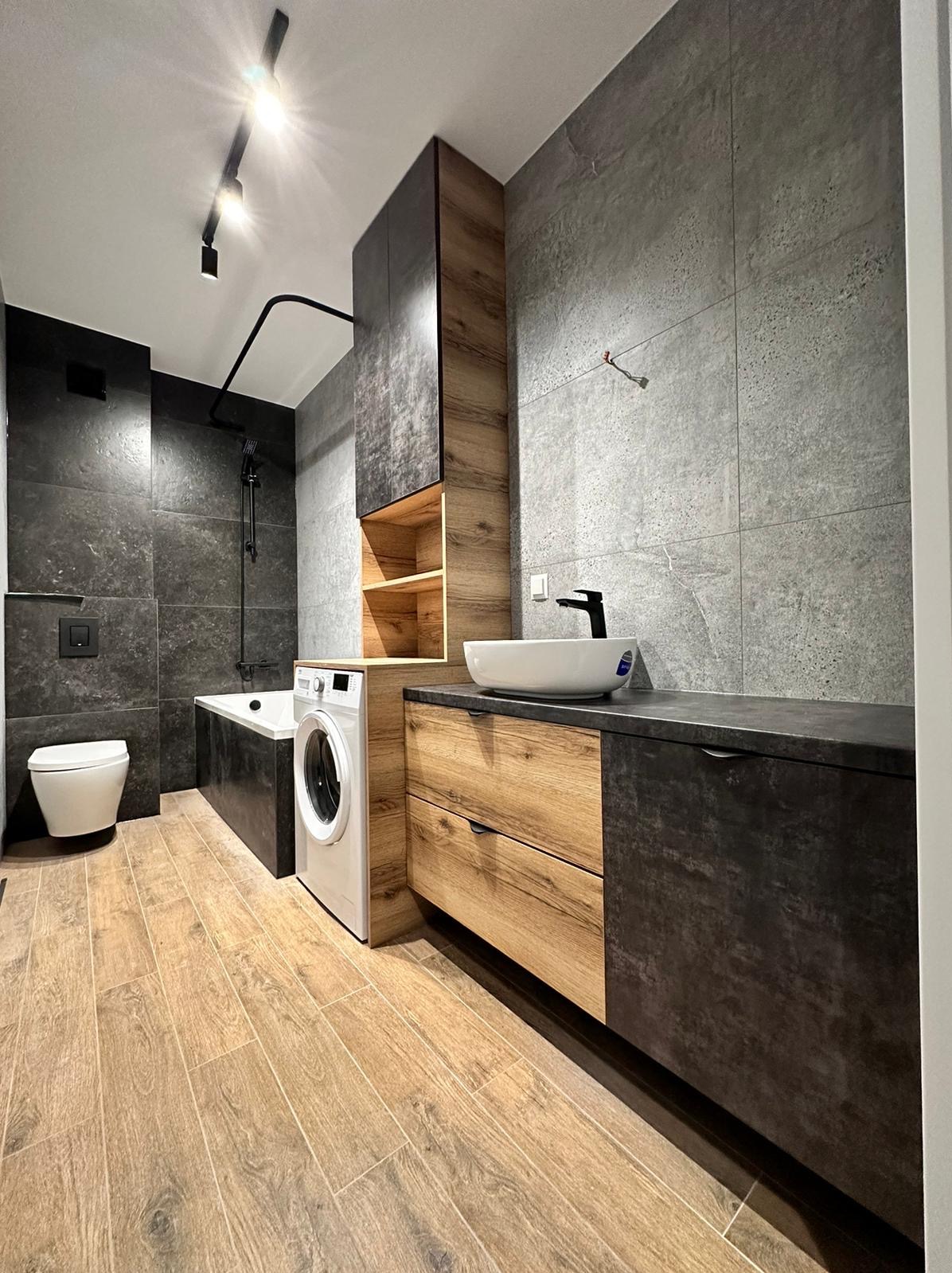 Nowoczesna łazienka w Toruniu z drewnianymi akcentami: szafka pod umywalkę z szufladami, wysoka szafka z otwartymi półkami, pralka obok wanny, szare płytki na ścianach i drewnopodobna podłoga.