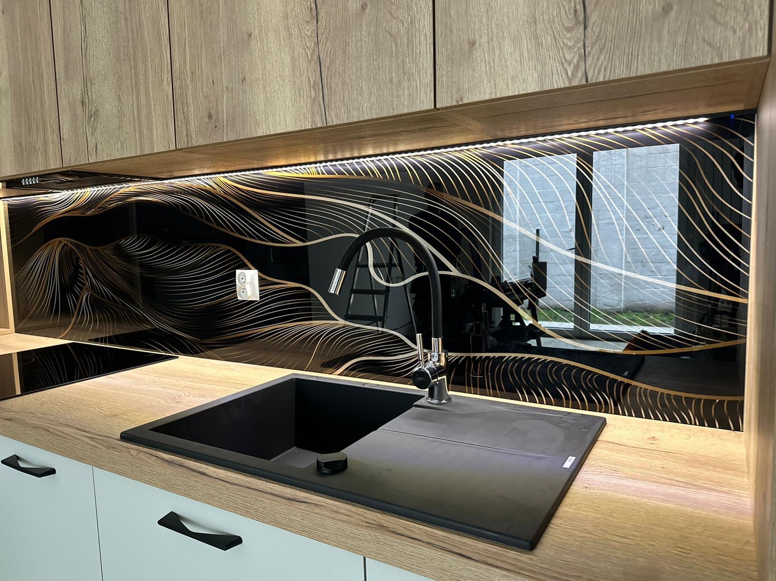 Czarny zlew z baterią na blacie z dekorem drewna, nad nim szklany panel z abstrakcyjnym złotym wzorem i oświetleniem LED, widoczne białe fronty szafek z czarnymi uchwytami.
