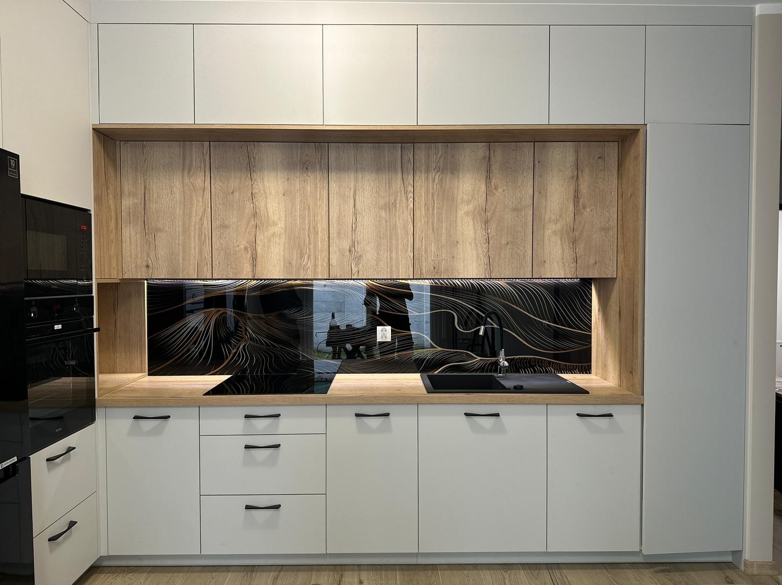 Nowoczesna kuchnia: białe szafki dolne z czarnymi uchwytami, drewnopodobne szafki górne, czarny szklany panel ścienny z złotymi wzorami, czarny zlew i płyta indukcyjna.