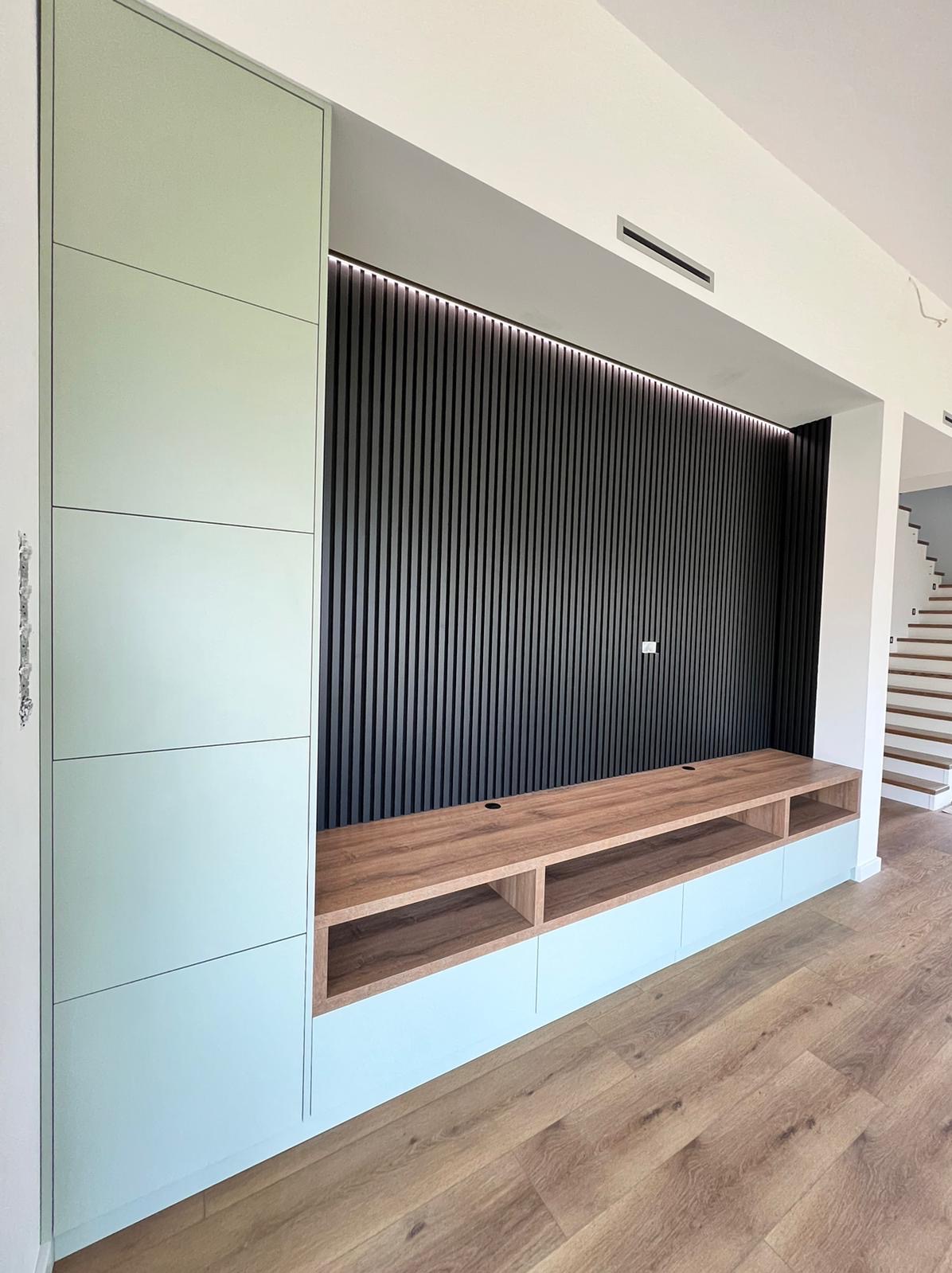 Nowoczesna zabudowa ściany telewizyjnej z pionowymi panelami dekoracyjnymi, szafką RTV z drewnianym blatem i szafką boczną w kolorze miętowym, na podłodze panele drewnopodobne.