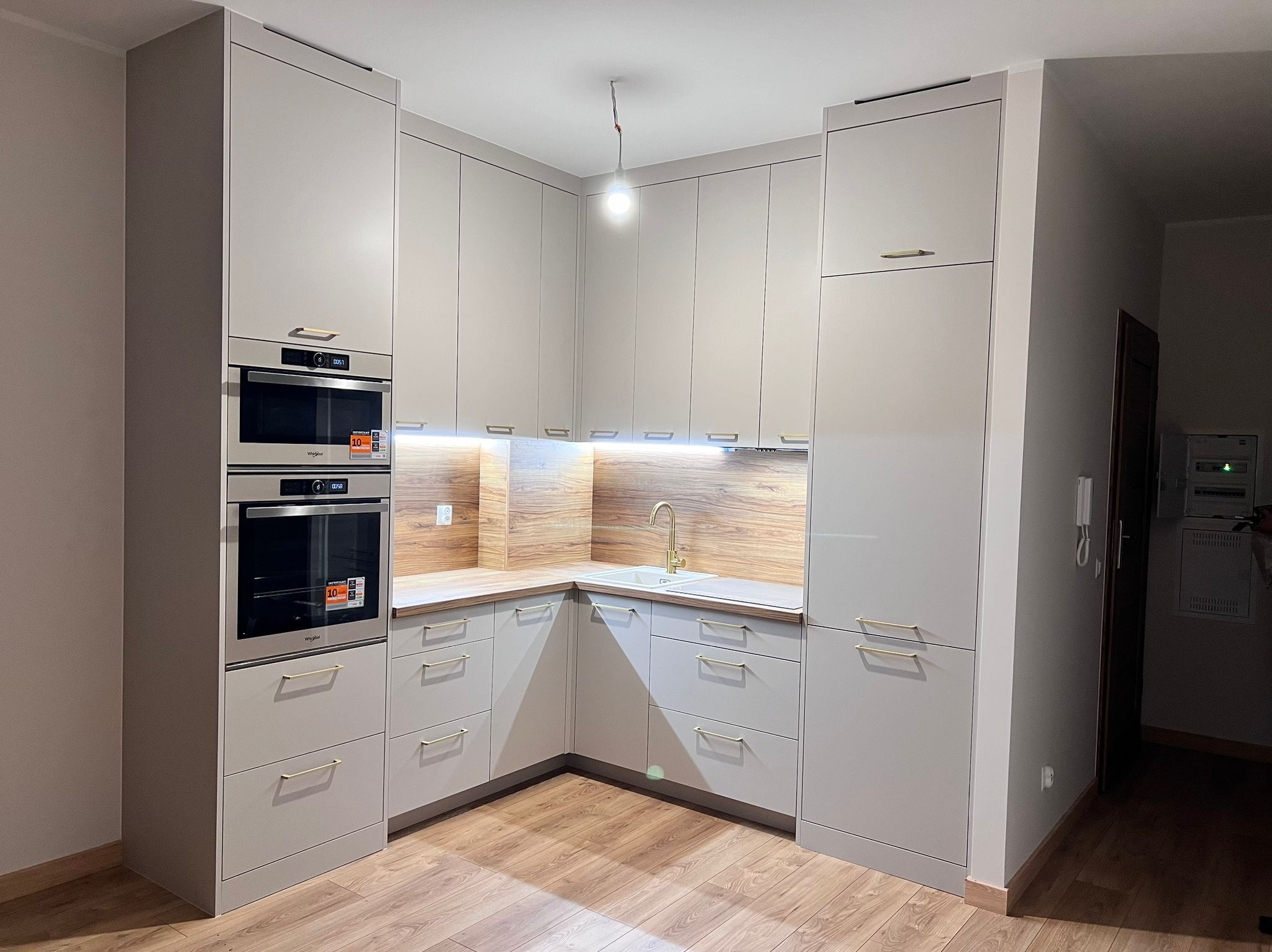 Jasnoszara kuchnia na wymiar z drewnianymi akcentami, złotymi uchwytami i wbudowanymi piekarnikami, oświetlona punktowo pod szafkami wiszącymi.