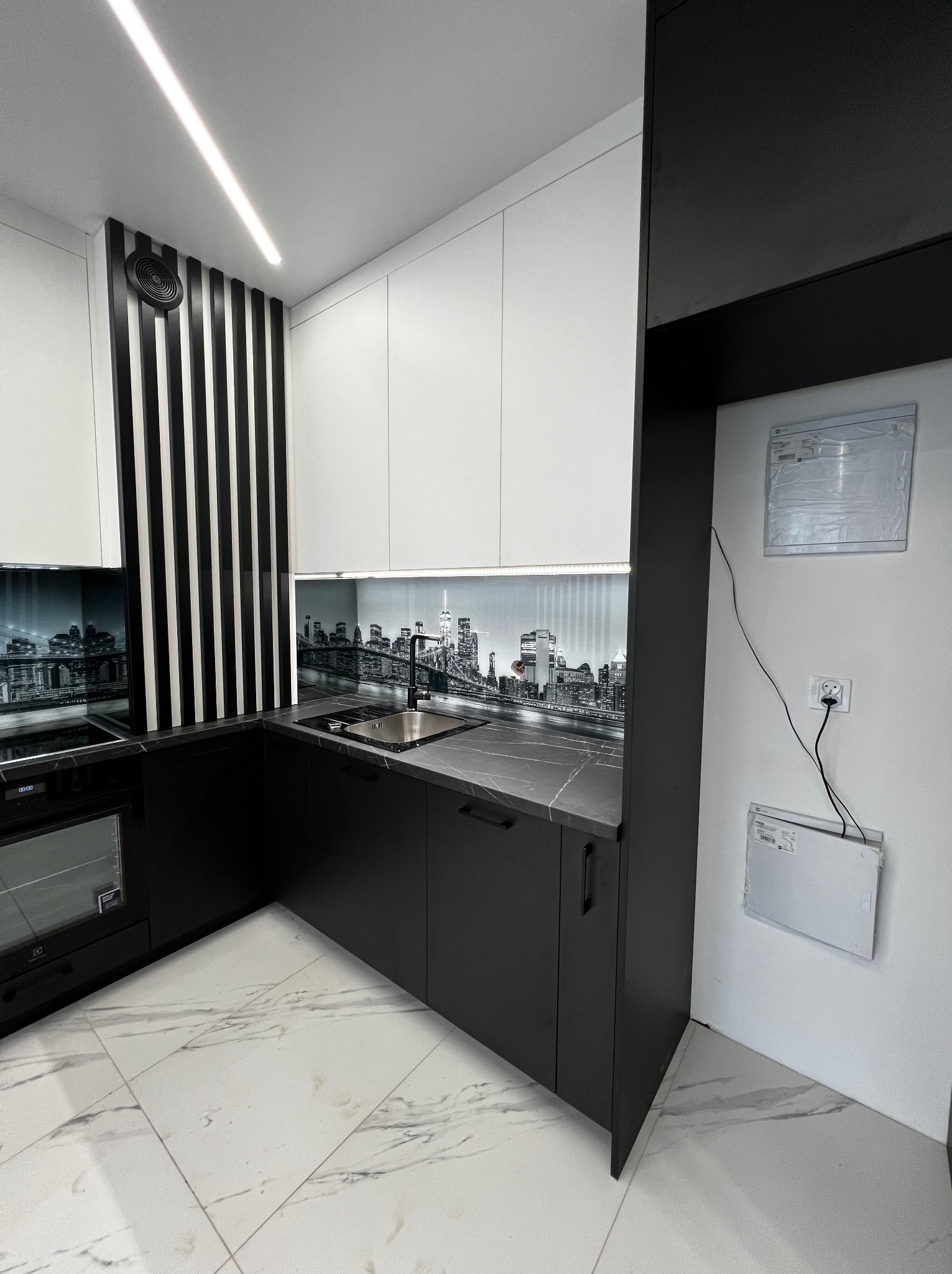 Nowoczesna kuchnia w czerni i bieli z blatem imitującym marmur i fototapetą z panoramą miasta nad zlewem, oświetlenie ledowe, białe szafki wiszące, czarne dolne szafki i pionowe czarno-białe panele...