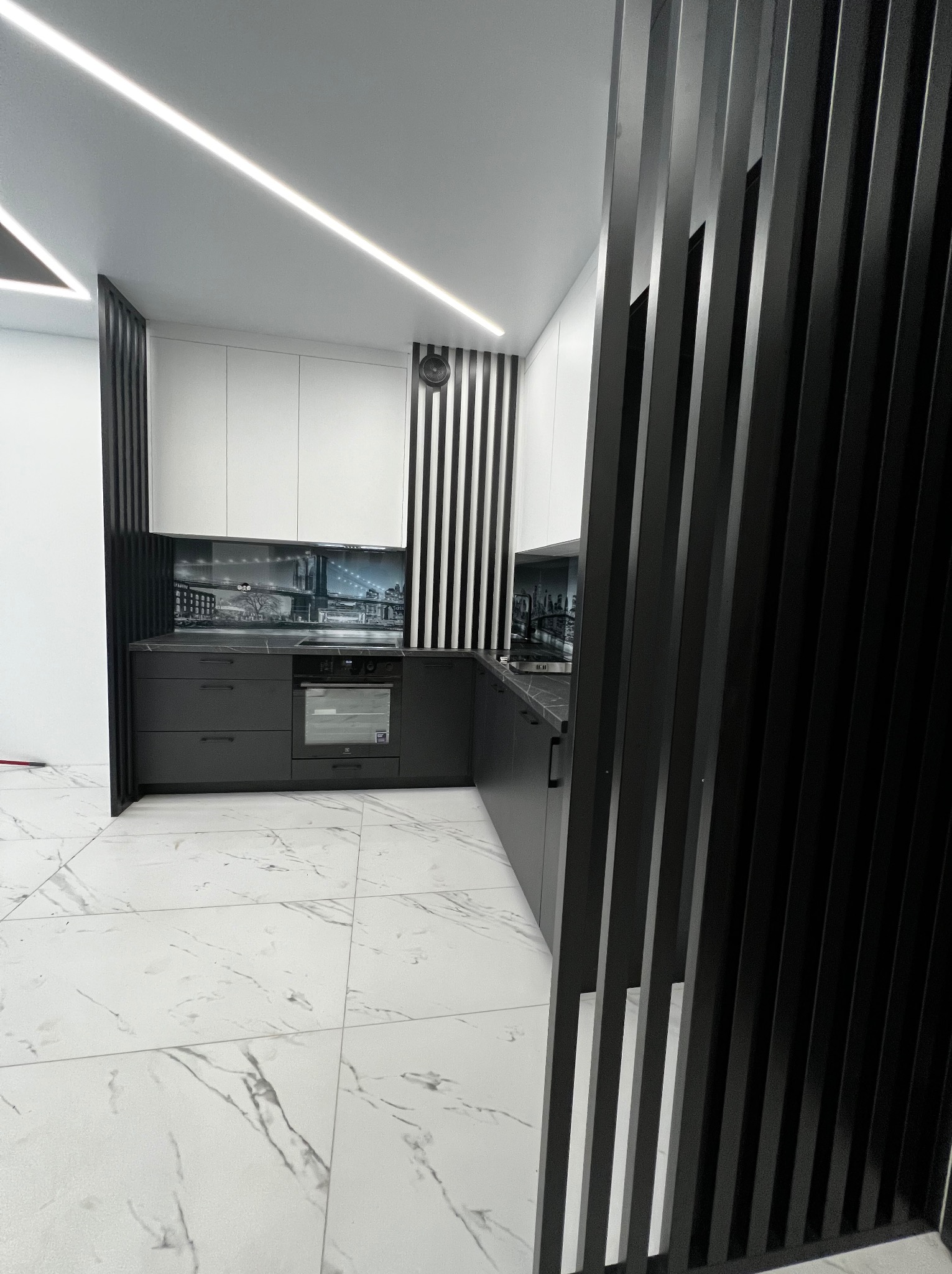 Nowoczesna kuchnia z czarnymi dolnymi szafkami i białymi górnymi, oddzielona od reszty pomieszczenia czarnymi, pionowymi listwami. Na ścianie nad blatem grafika z motywem miejskim.