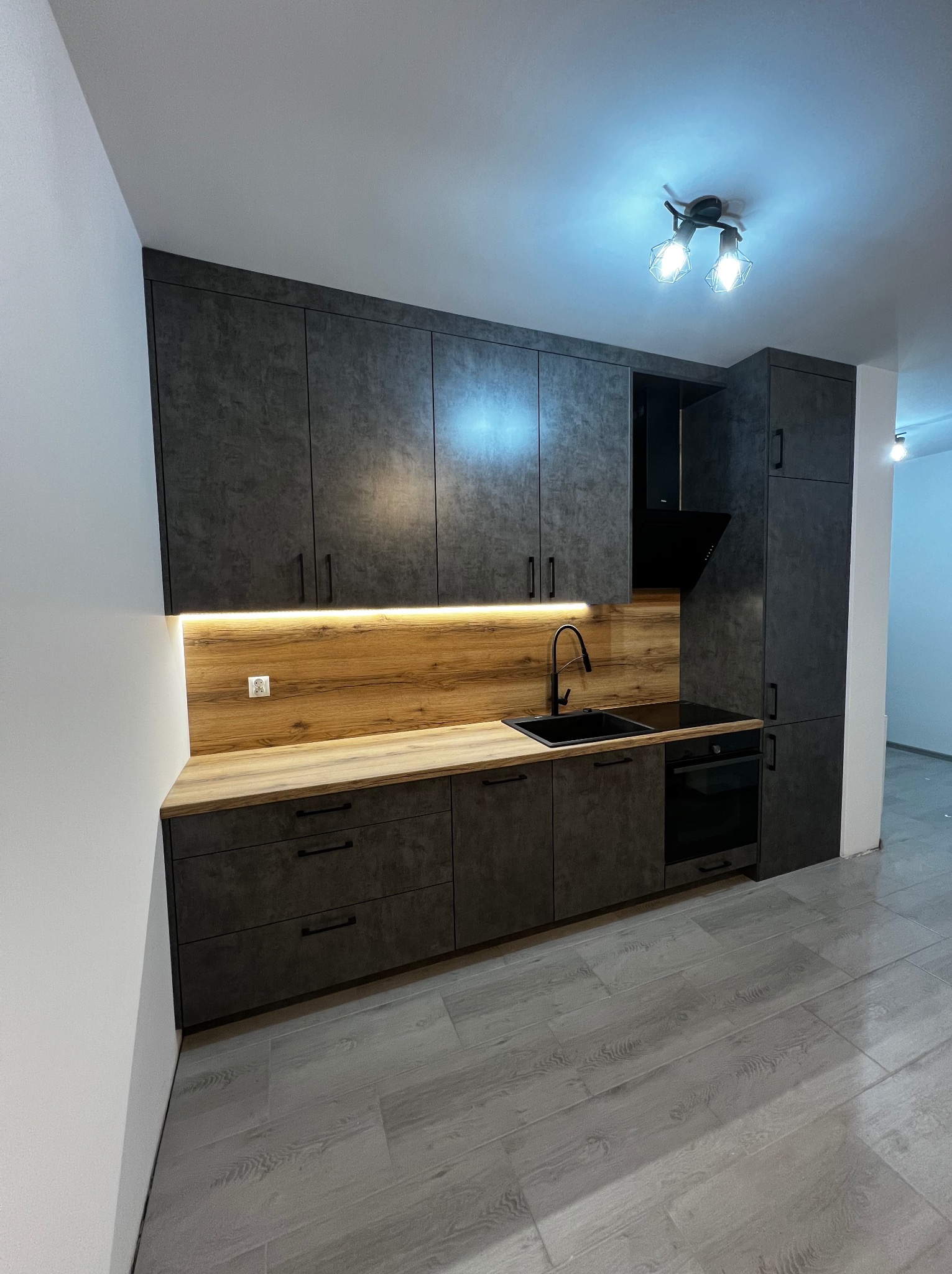 Nowoczesna kuchnia w odcieniach szarości z drewnianym blatem, oświetleniem LED podszafkowym i czarną armaturą, minimalistyczny design z frontami bezuchwytowymi i szarą podłogą imitującą drewno.