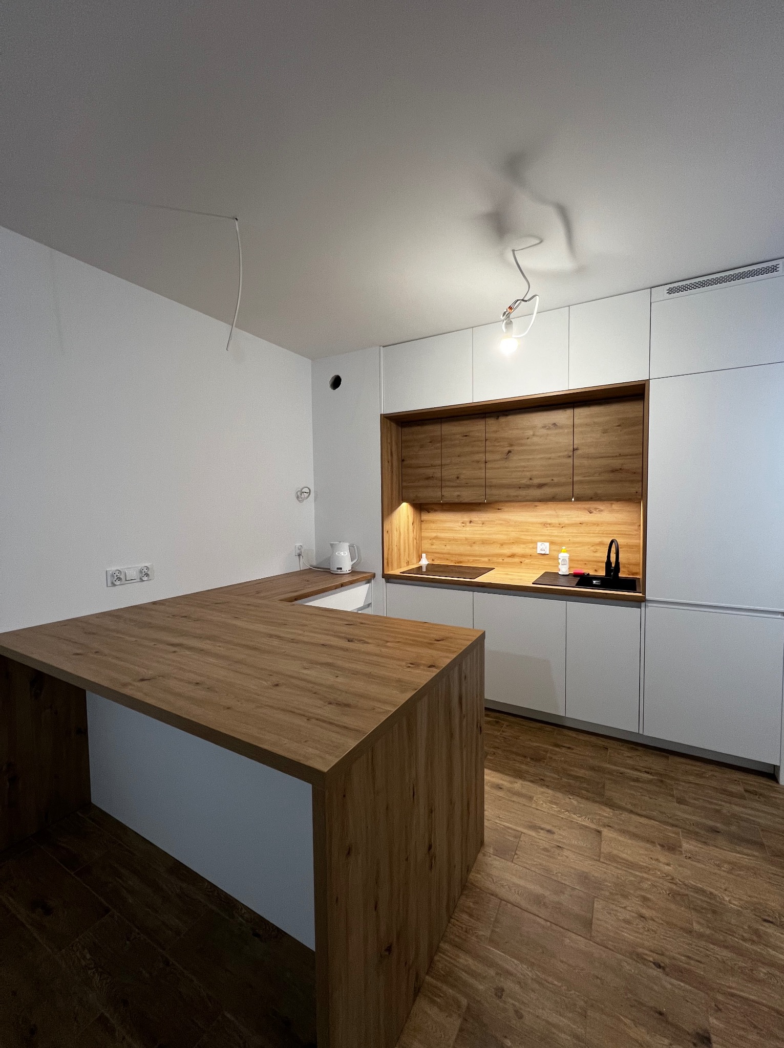 Nowoczesna kuchnia z białymi szafkami i drewnianym blatem wyspowym; widoczny fragment kabla zwisającego z sufitu.