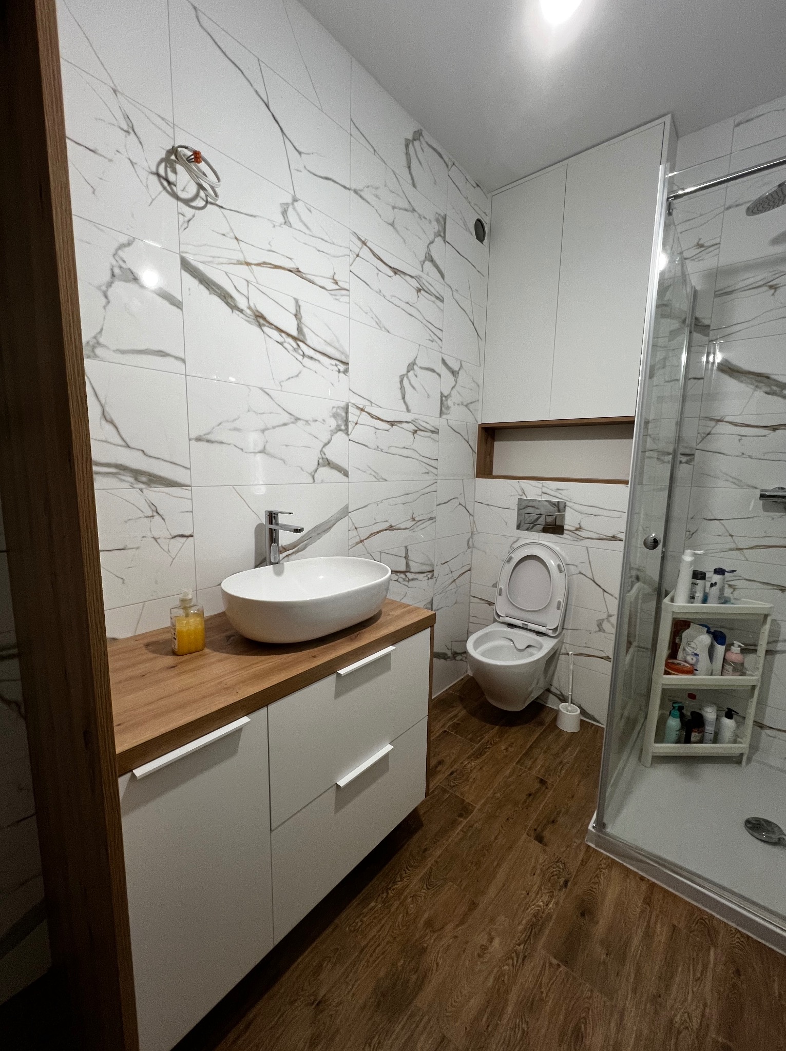 Nowoczesna łazienka z blatem drewnopodobnym pod umywalką nablatową, białymi szafkami, marmurowymi płytkami na ścianach i kabiną prysznicową.