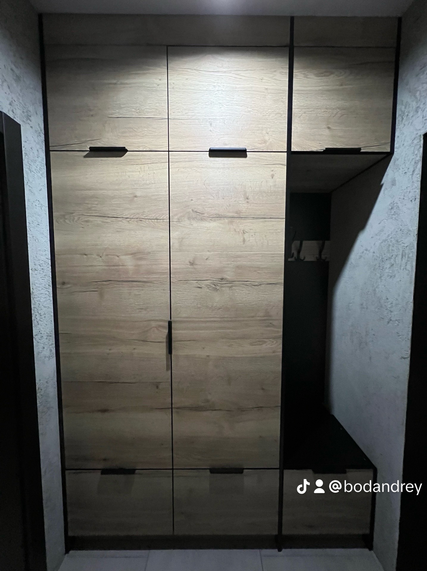 Szafa wnękowa z drewnopodobnymi frontami i czarnymi uchwytami, z otwartą wnęką na buty i wieszaki, umieszczona w korytarzu o szarych ścianach.