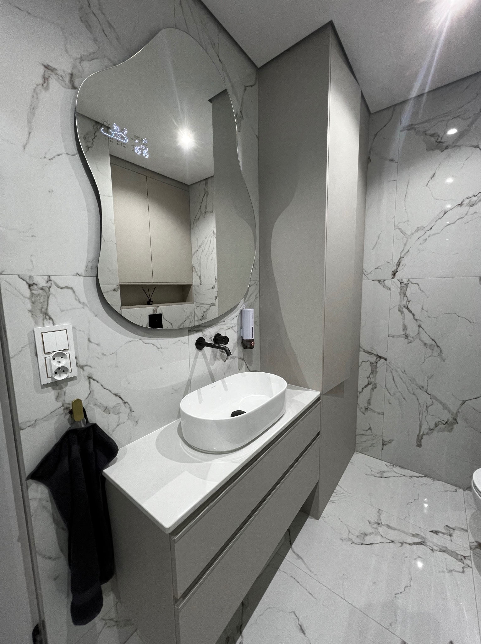 Nowoczesna łazienka z umywalką nablatową, szafką pod umywalką w kolorze szarym, wysoką szafką, lustrem o nieregularnym kształcie z wyświetlaczem oraz marmurowymi płytkami na ścianach i podłodze.