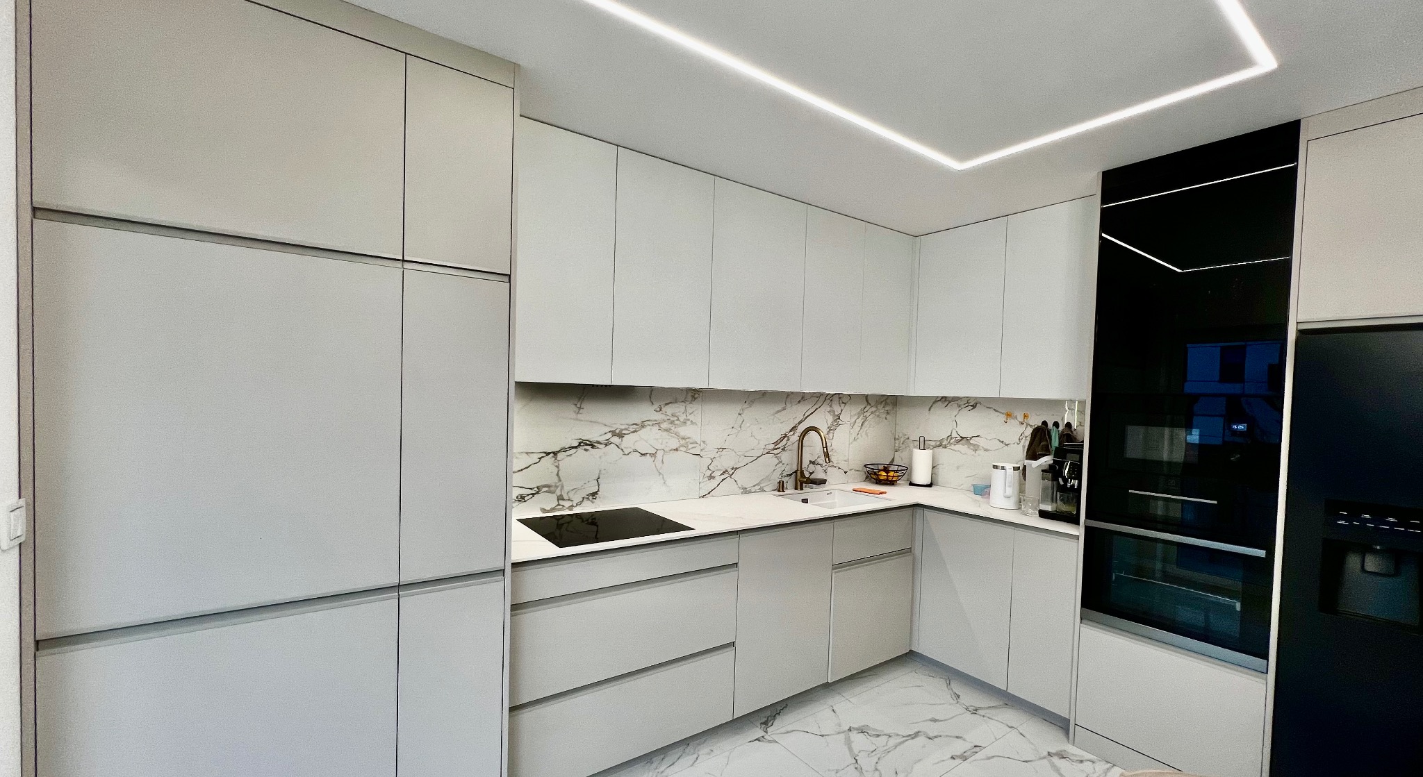 Nowoczesna, minimalistyczna kuchnia w odcieniach szarości z marmurowym blatem i podłogą, oświetleniem LED na suficie oraz czarnymi sprzętami AGD.