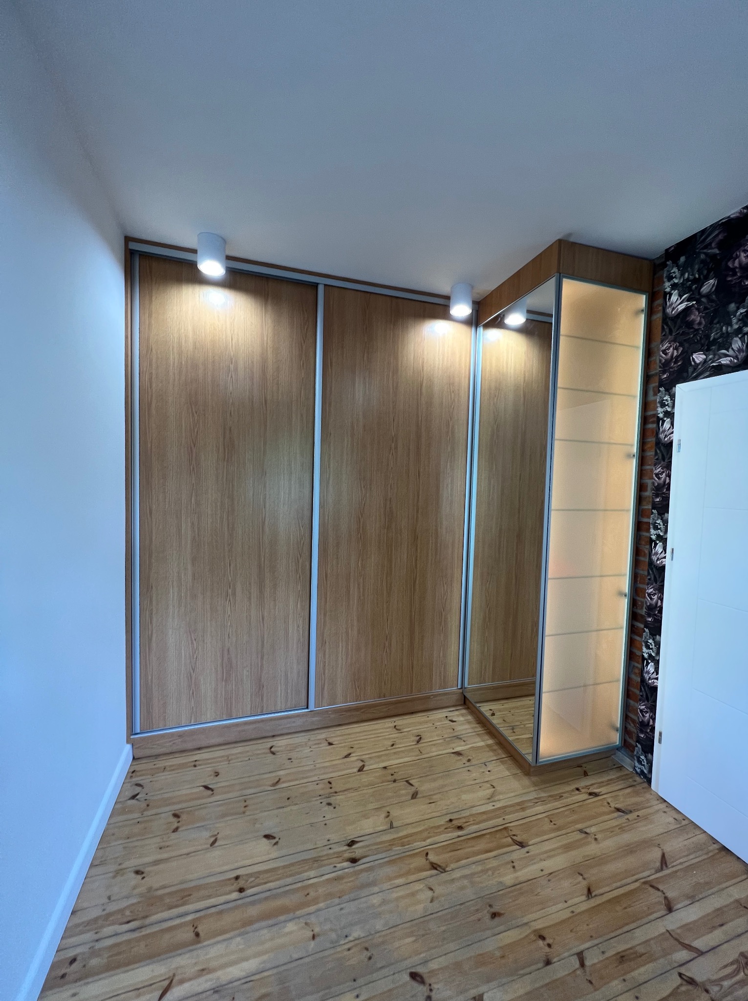 Wnętrze pokoju z dużą szafą przesuwną z drewnopodobnymi frontami i przeszklonym segmentem z półkami, oświetloną od góry, na drewnianej podłodze.