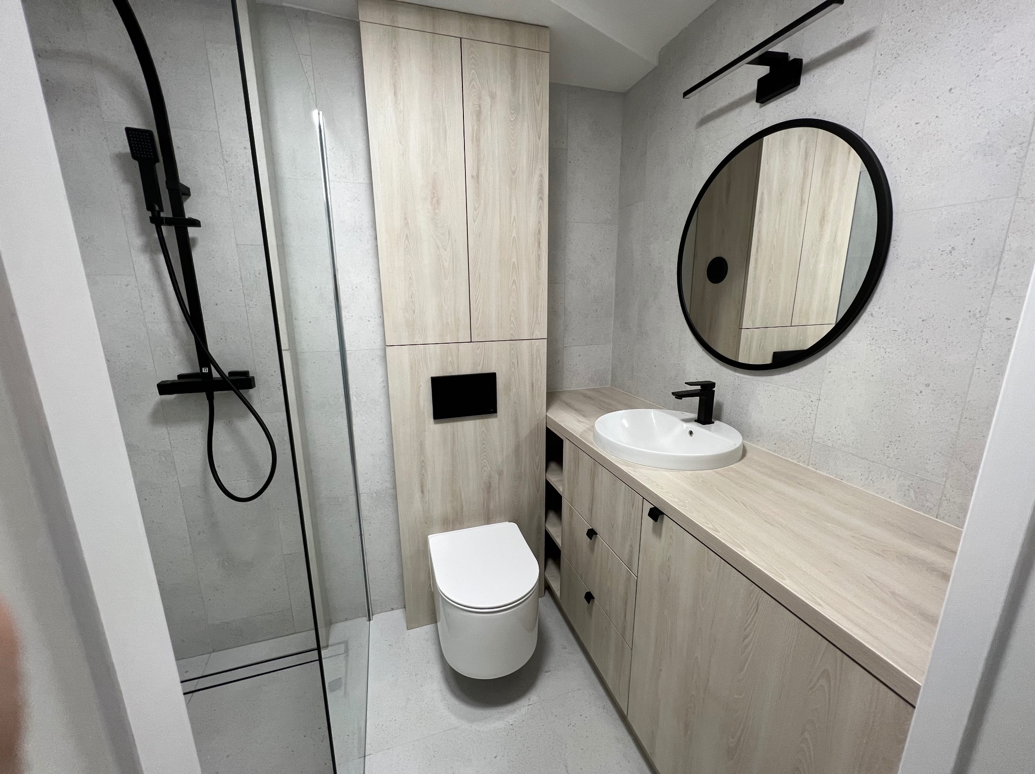 Nowoczesna łazienka z kabiną prysznicową, wiszącą toaletą i szafką pod umywalką w kolorze jasnego drewna, uzupełniona czarnymi akcentami.
