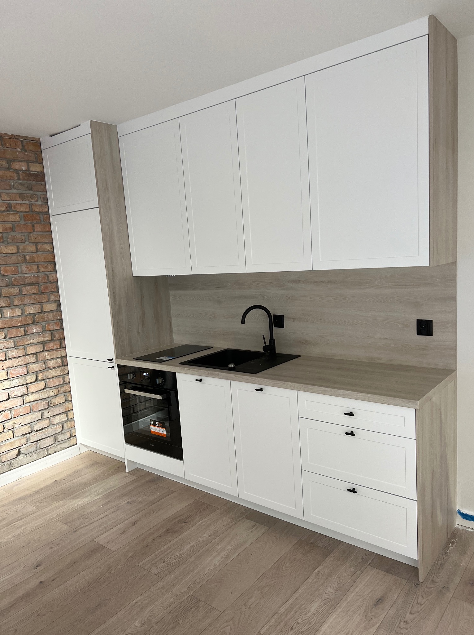 Nowoczesna kuchnia z białymi frontami, drewnopodobnym blatem i czarną armaturą, kontrastująca z ceglaną ścianą w tle.