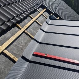 Pokrycia Dachowe Jantar - Częściowo pokryty dach: czarne dachówki ceramiczne i szare panele blaszane, poziomica spoczywająca na panelach, widoczne łaty drewniane i membrana dachowa.