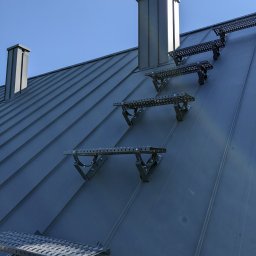 Pokrycia Dachowe Jantar - Metalowy dach z zamontowanymi stopniami drabinkowymi i kominami w kolorze grafitowym, widziany pod kątem z dołu w słoneczny dzień.