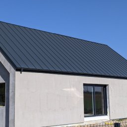 Pokrycia Dachowe Jantar - Nowoczesny dom w stanie surowym zamkniętym z grafitowym dachem z blachy na rąbek stojący, widoczne rynny i obróbki blacharskie, okna z czarnymi ramami, błękitne niebo w tle.