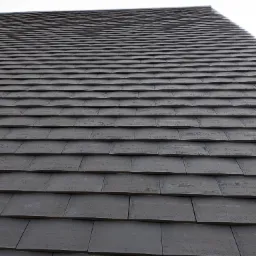 Ciemnoszara dachówka ceramiczna ułożona w regularny wzór, widok z dołu pod kątem, fragment dachu na tle jasnego nieba.
