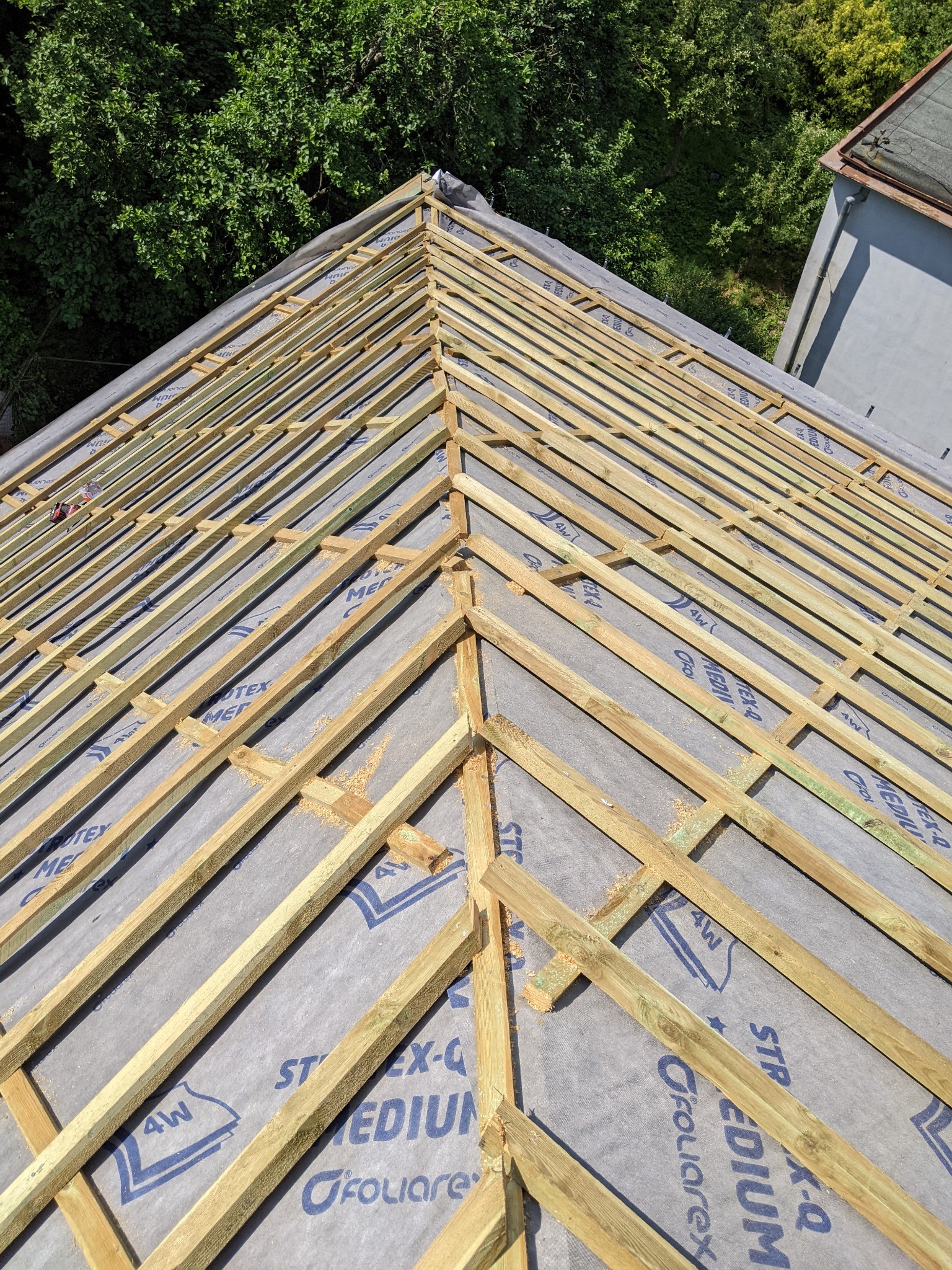 Konstrukcja drewniana dachu z łatami na membranie, widok z góry, w tle zieleń drzew i fragment budynku.