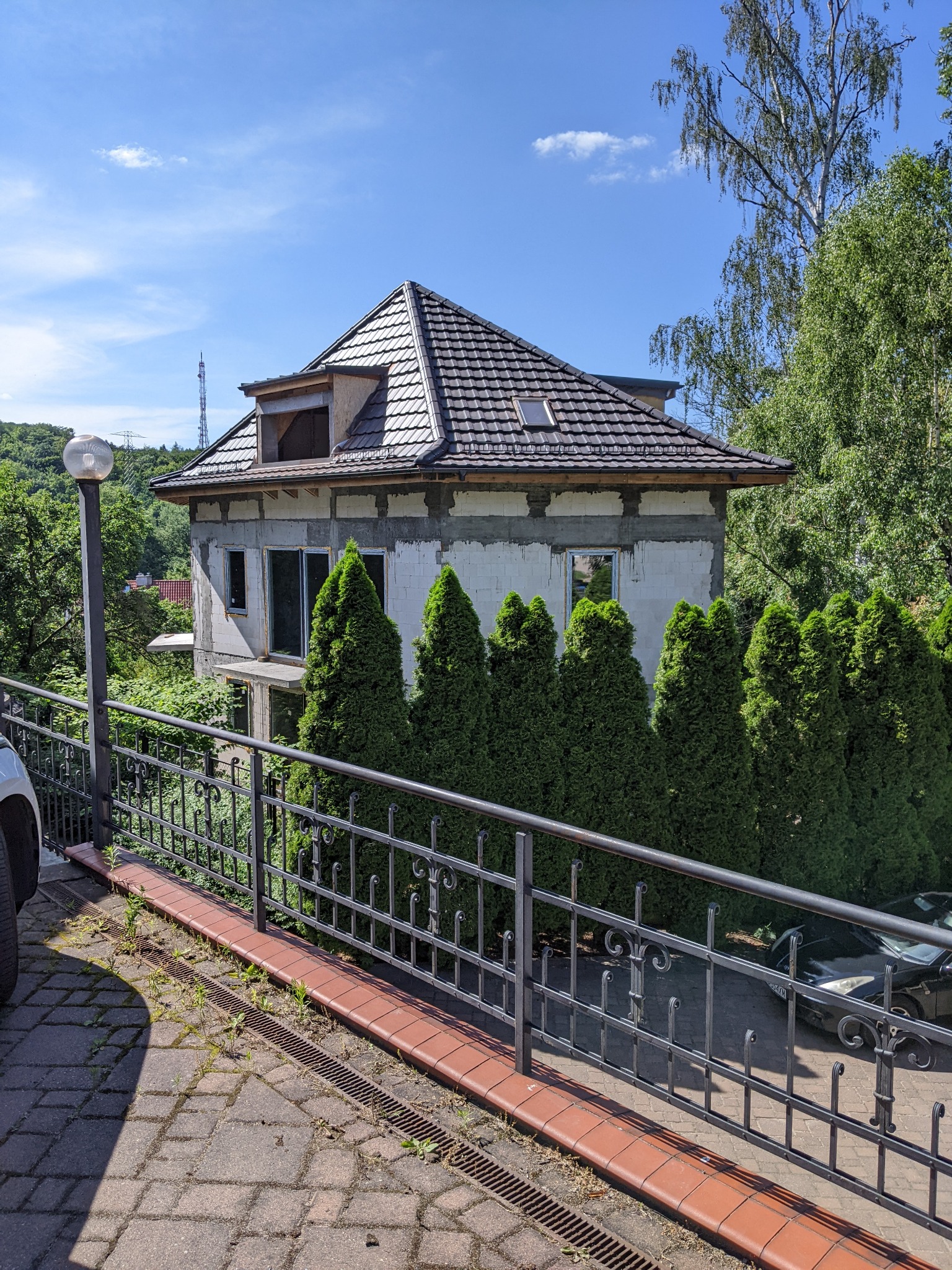 Niedokończony budynek z ciemnym dachem z dachówki ceramicznej, widoczny zza ozdobnego metalowego ogrodzenia i rzędu wysokich, stożkowych krzewów.