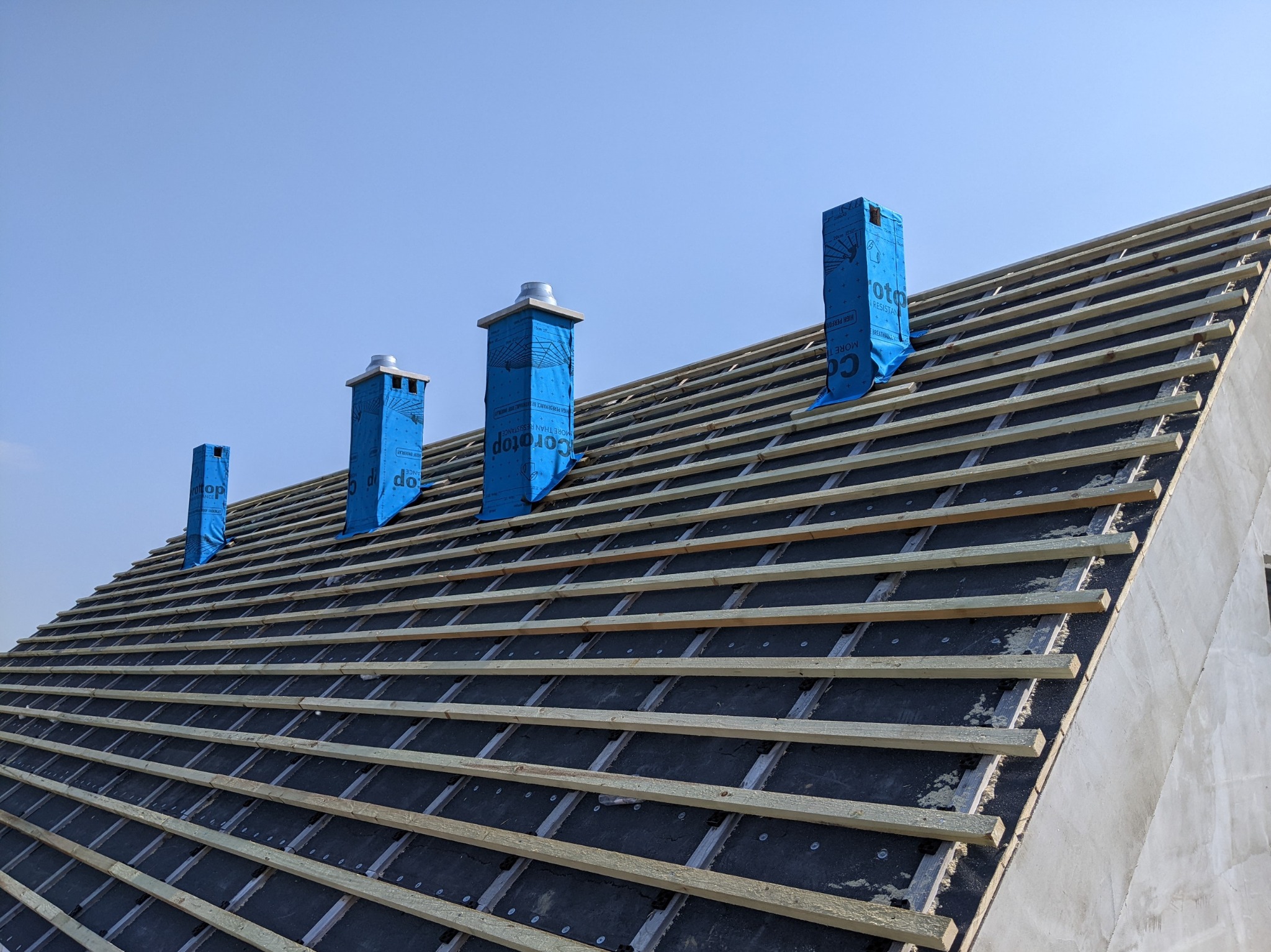 Konstrukcja dachu z łatami i kontrłatami, zabezpieczone kominy pokryte niebieską folią ochronną z nadrukiem, widok z dołu na tle błękitnego nieba.