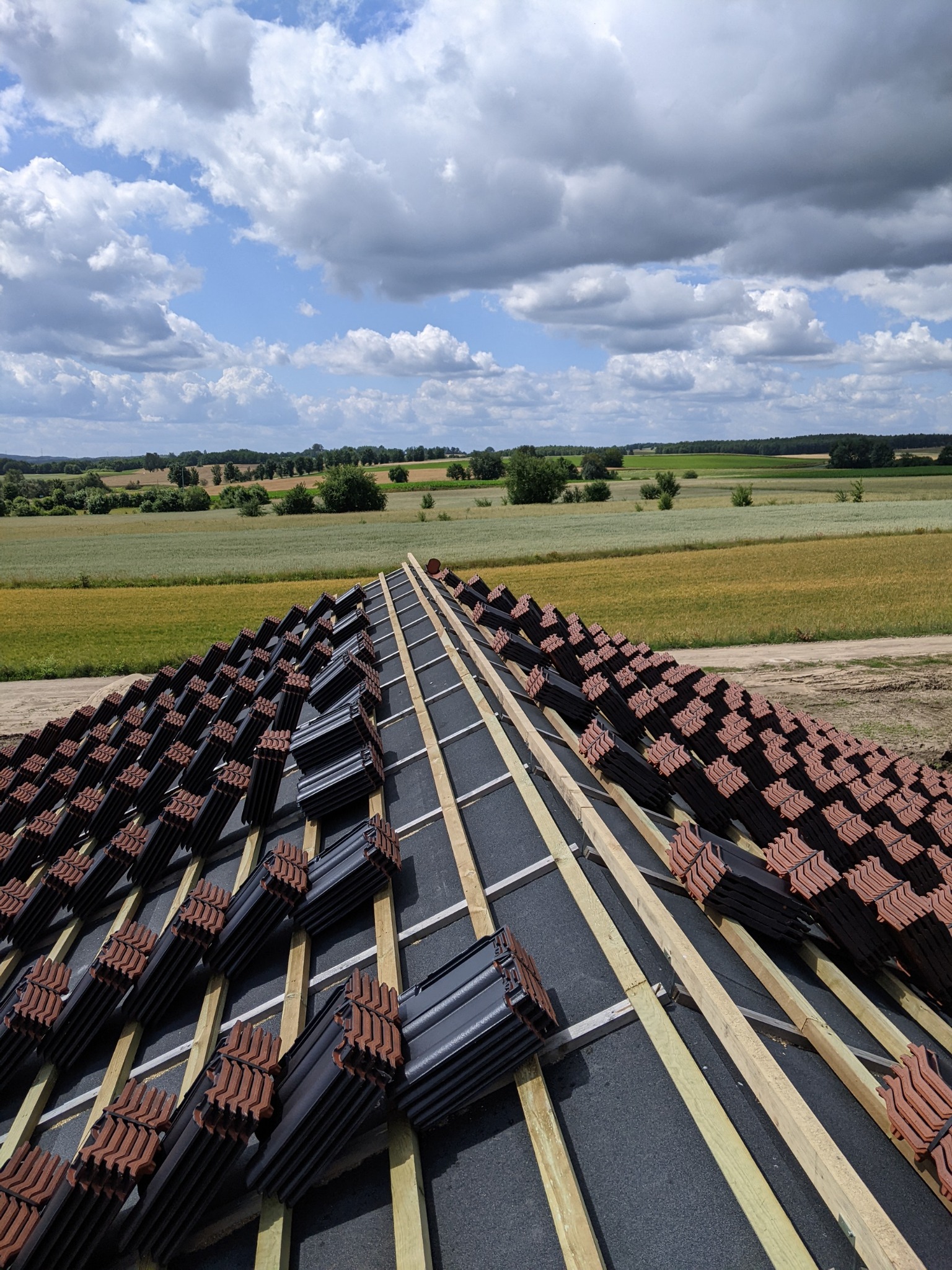 Przygotowanie do ułożenia dachówki ceramicznej w kolorze grafitowym z brązowymi krawędziami na łatach dachowych z widokiem na pola uprawne pod błękitnym, pochmurnym niebem.