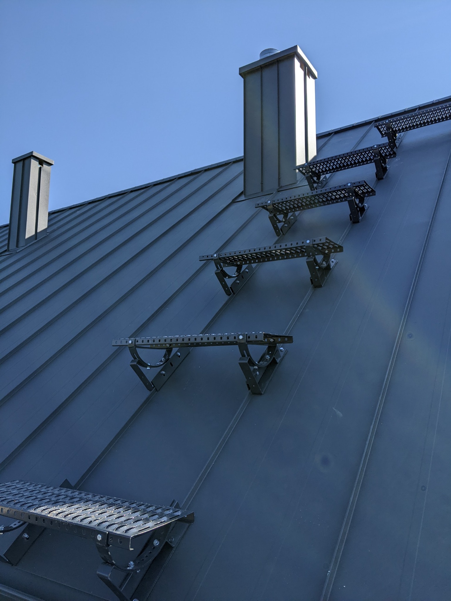 Metalowy dach z zamontowanymi stopniami drabinkowymi i kominami w kolorze grafitowym, widziany pod kątem z dołu w słoneczny dzień.