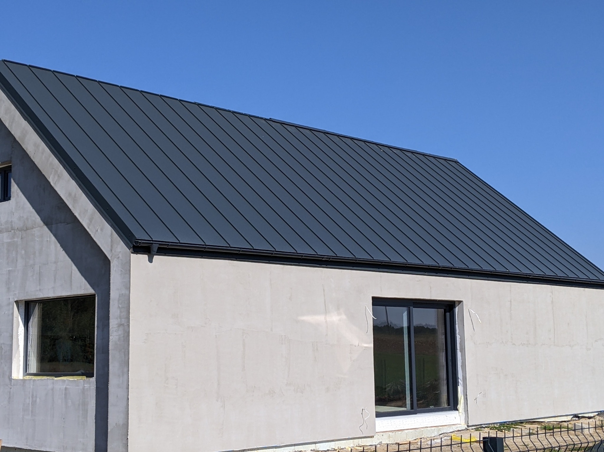 Nowoczesny dom w stanie surowym zamkniętym z grafitowym dachem z blachy na rąbek stojący, widoczne rynny i obróbki blacharskie, okna z czarnymi ramami, błękitne niebo w tle.