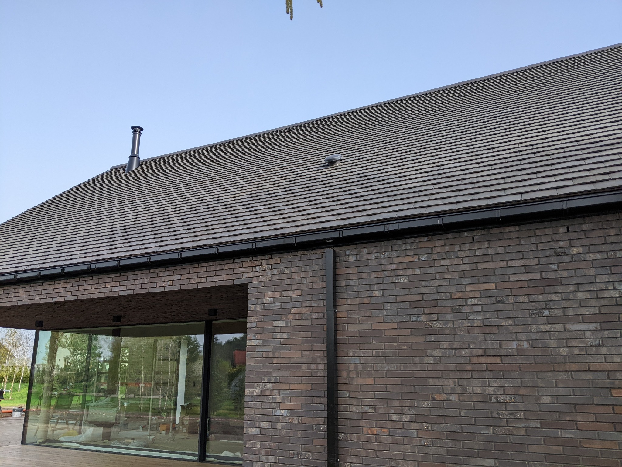 Nowoczesny dom z ciemnej cegły z dachem krytym dachówką ceramiczną w odcieniach szarości, widoczne rynny w kolorze czarnym i komin.