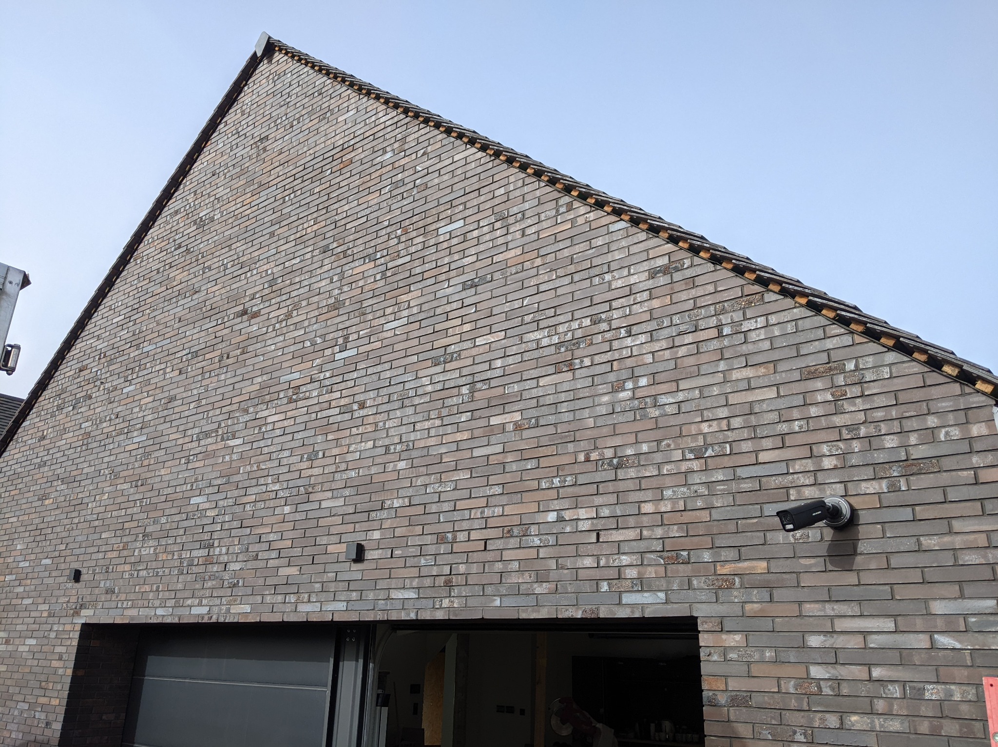 Ściana szczytowa budynku mieszkalnego wykończona klinkierem w odcieniach szarości i brązu, widoczna kamera monitoringu, brama garażowa i fragment dachu z wystającymi elementami okapu.