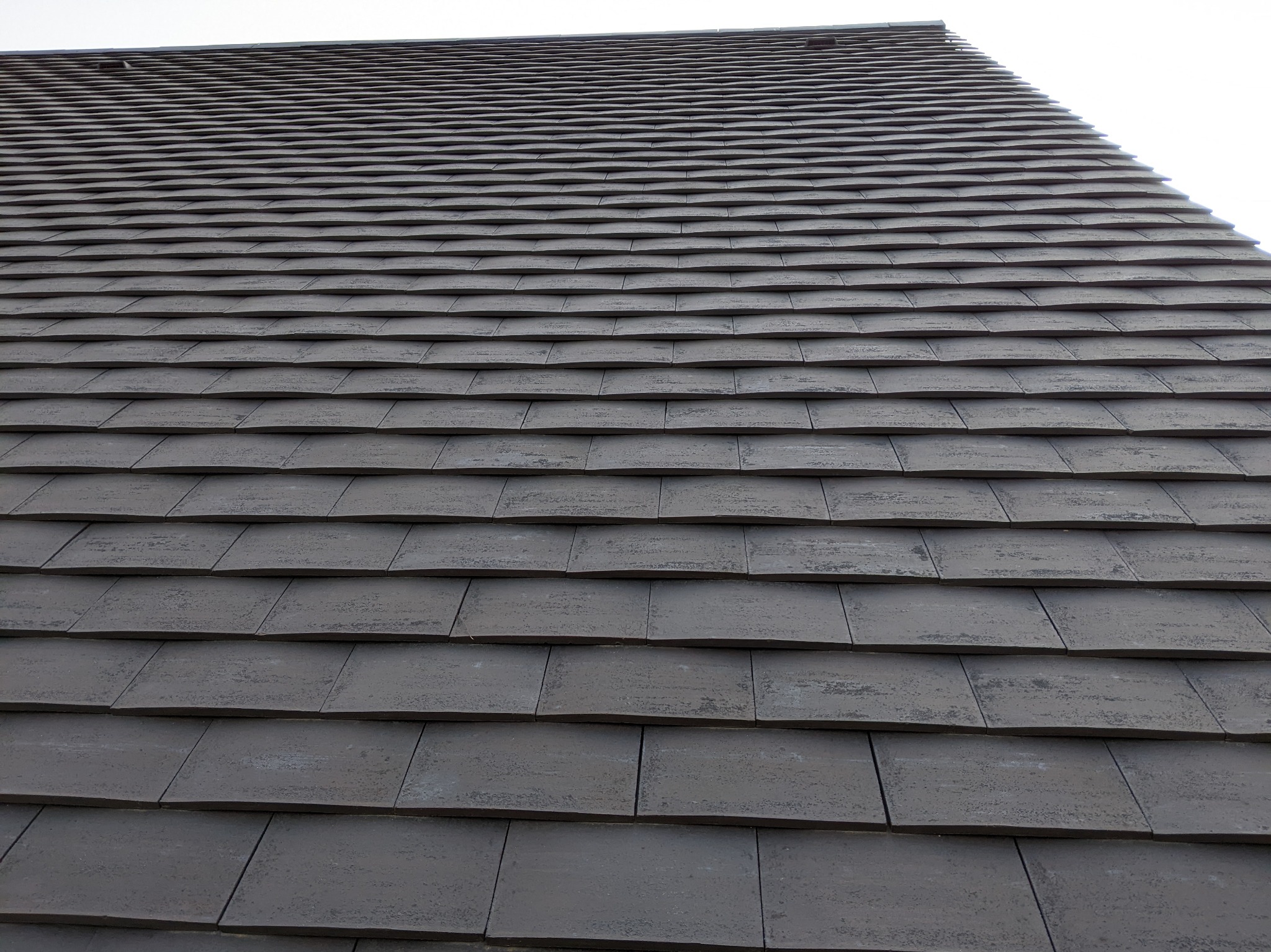 Ciemnoszara dachówka ceramiczna ułożona w regularny wzór, widok z dołu pod kątem, fragment dachu na tle jasnego nieba.