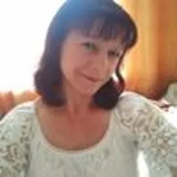 Portret kobiety z krótkimi brązowymi włosami i grzywką, ubranej w białą bluzkę z koronkowymi rękawami, uśmiechającej się w słonecznym świetle.