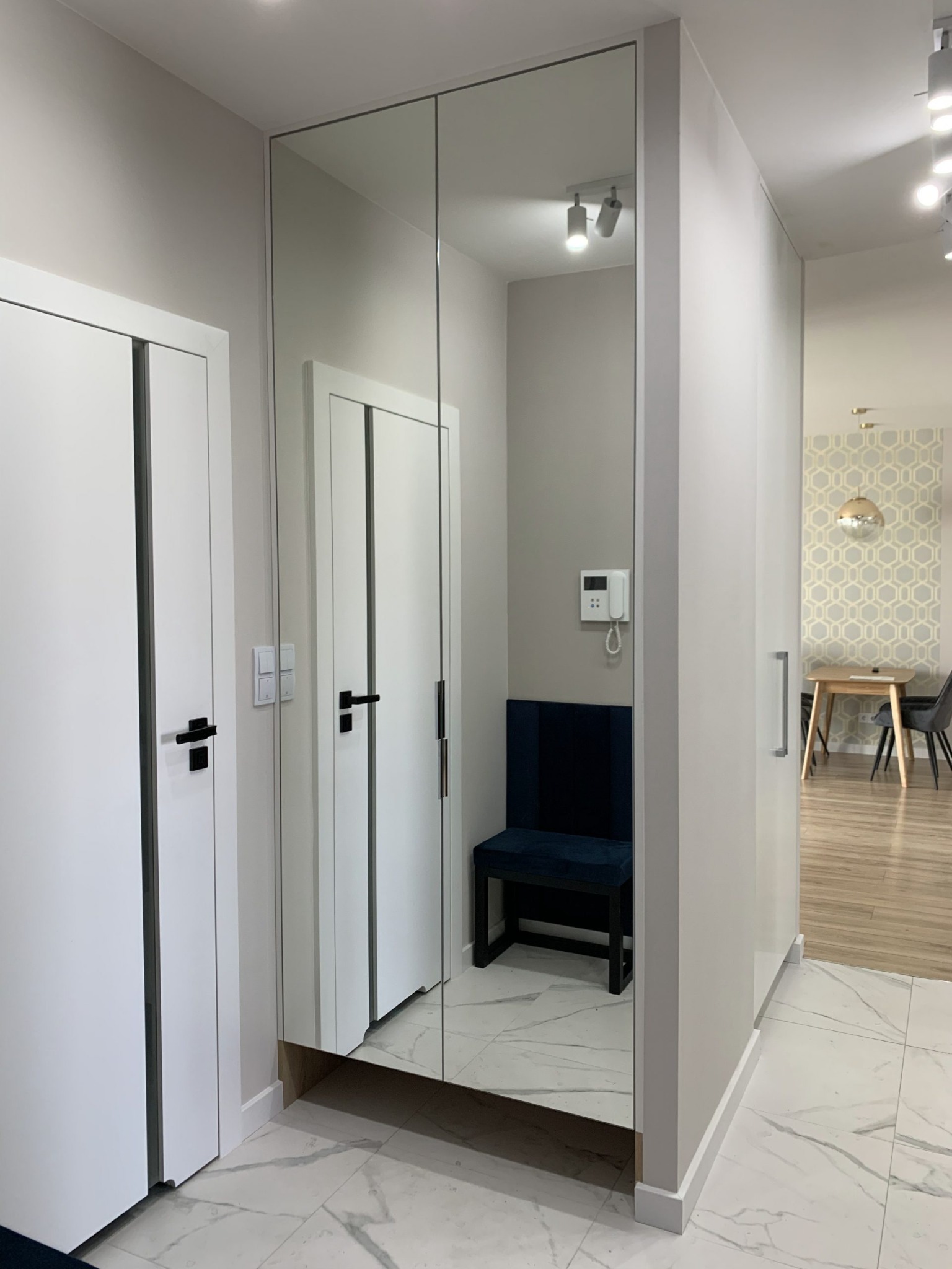 Przedpokój z zabudową lustrzaną na wymiar, ukrywającą siedzisko z granatową tapicerką, białe drzwi po lewej, marmurowa podłoga, widok na jadalnię w tle.
