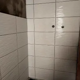 Ściana w łazience wyłożona białymi, połyskującymi płytkami z delikatnym falistym wzorem. Widoczne dwa punkty instalacji wodnej oraz surowy fragment ściany w rogu.