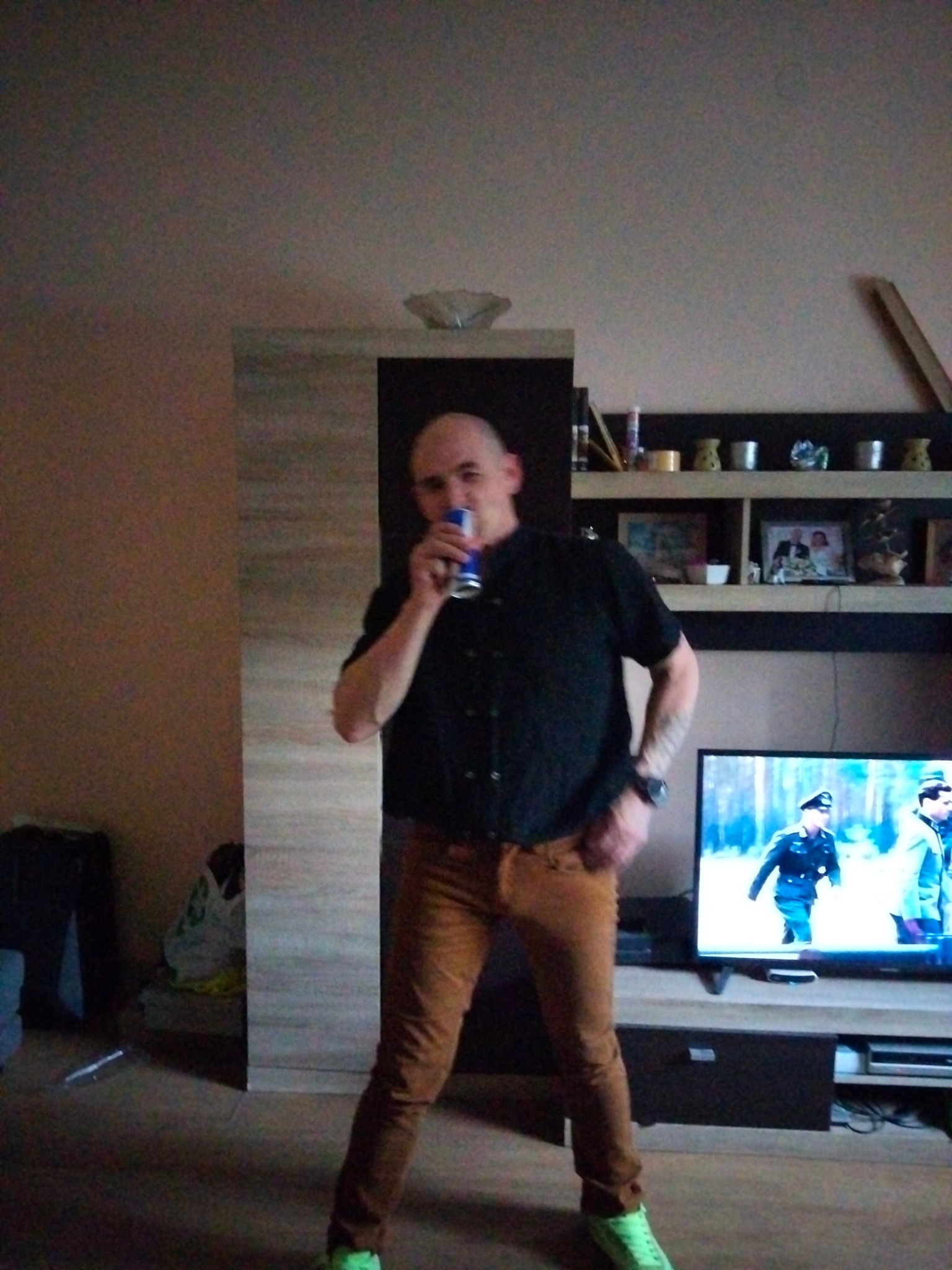 Mężczyzna pije napój energetyczny w salonie z telewizorem wyświetlającym film wojenny, obok szafka z półkami i pamiątkami, w tle ściana w odcieniach beżu.