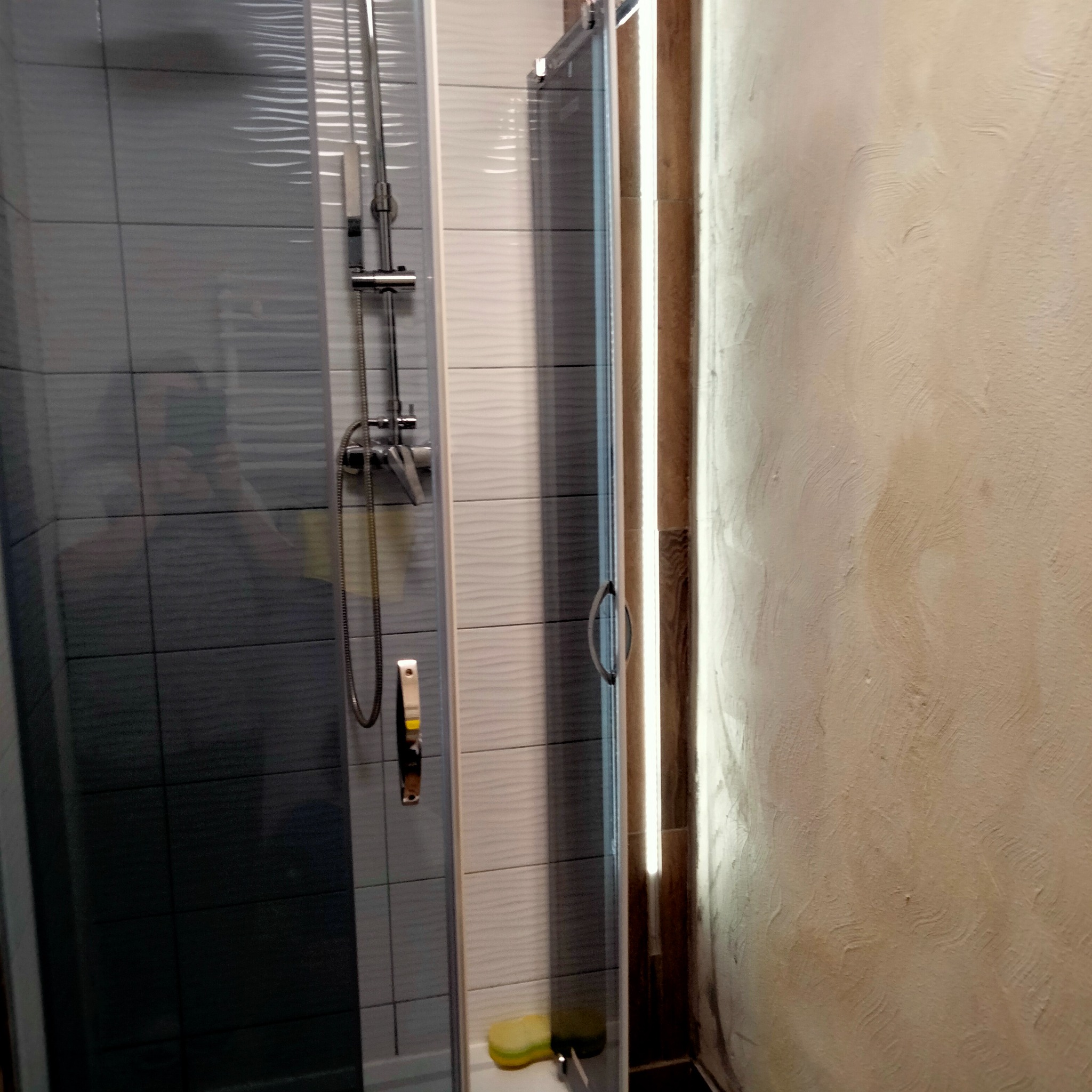 Wykończona kabina prysznicowa z szarymi i białymi płytkami o falistej strukturze, widoczna armatura i gąbka w rogu, ściana obok kabiny z surowym tynkiem.