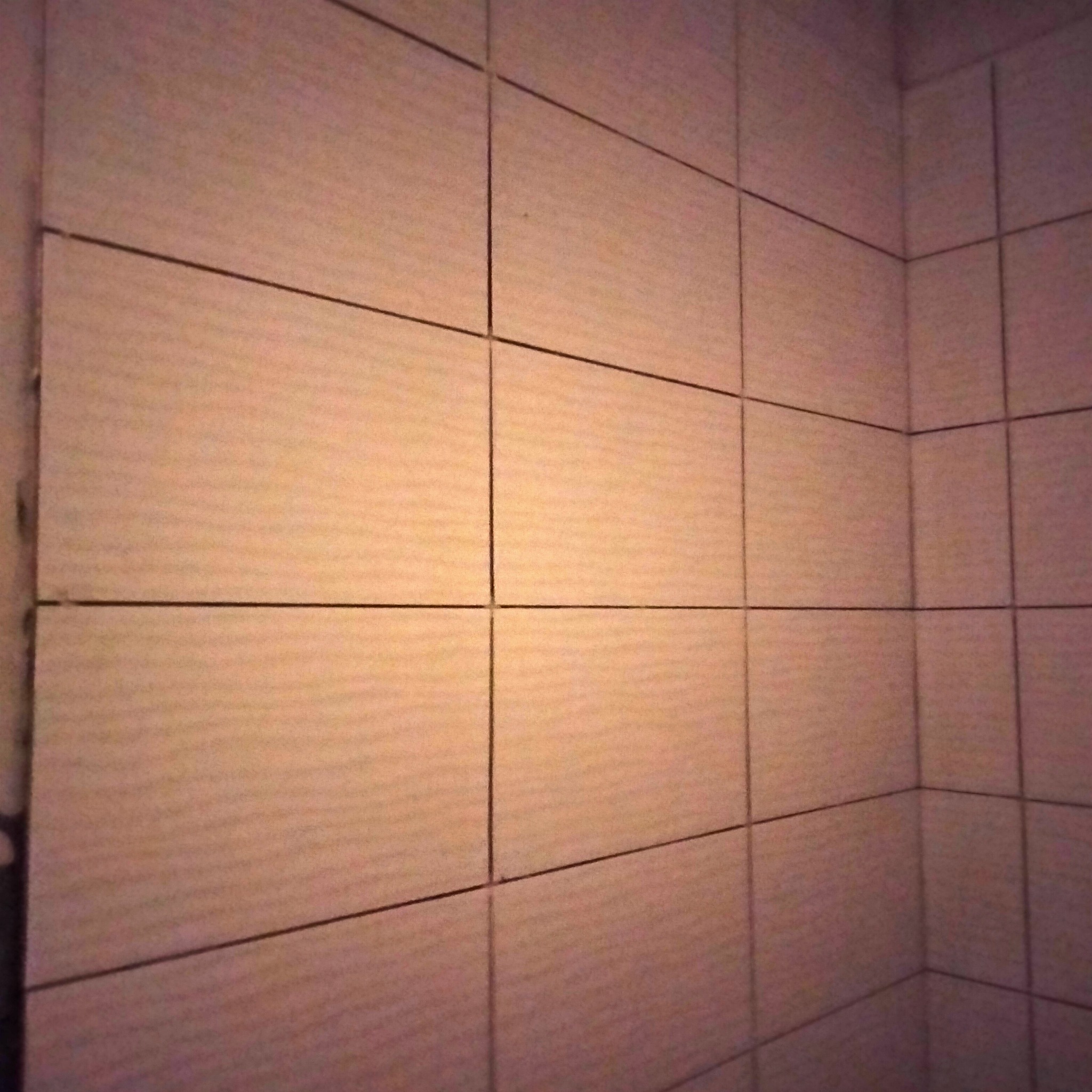 Wyłożona jasnymi, kwadratowymi płytkami ceramicznymi ściana w rogu pomieszczenia; widoczne ciemne fugi i minimalne niedociągnięcia przy krawędzi.