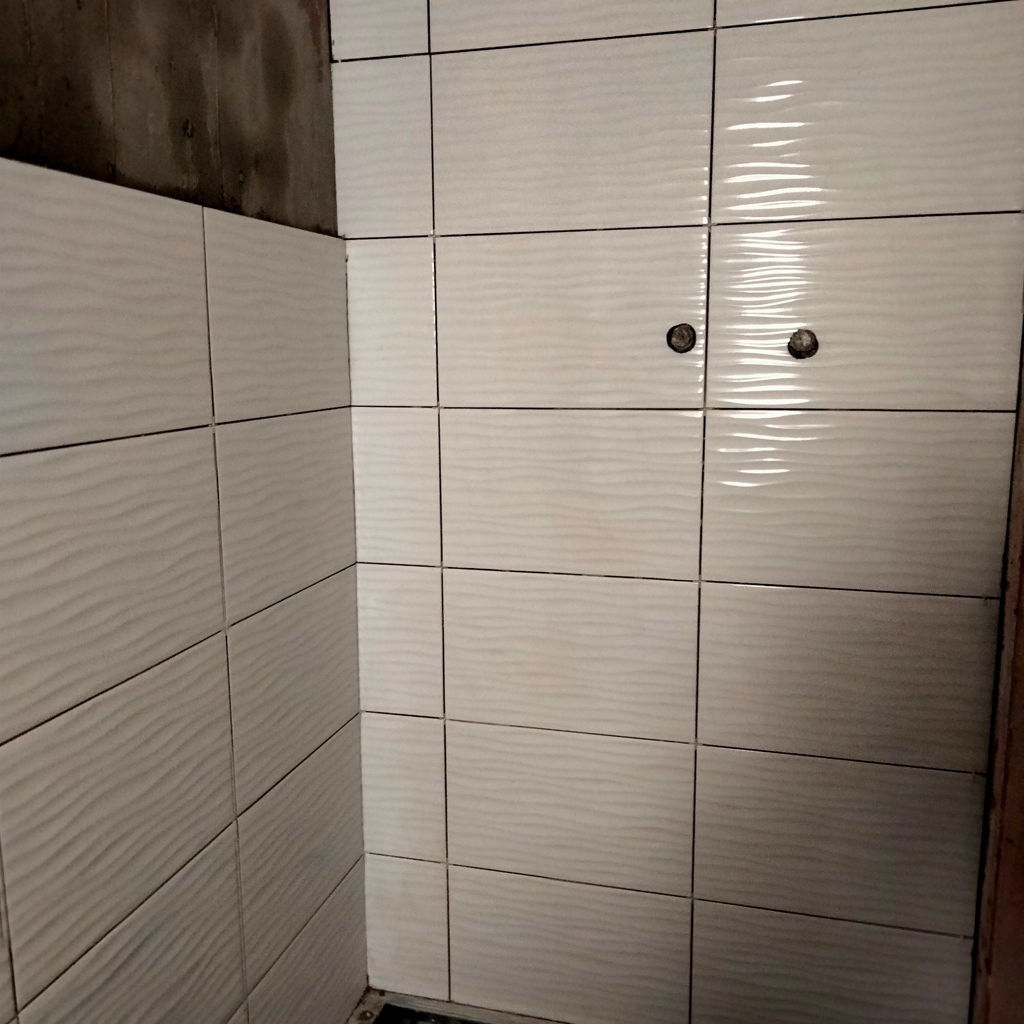 Ściana w łazience wyłożona białymi, połyskującymi płytkami z delikatnym falistym wzorem. Widoczne dwa punkty instalacji wodnej oraz surowy fragment ściany w rogu.