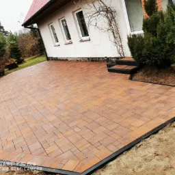 Nowo ułożony, geometryczny wz&oacute;r z brązowej kostki brukowej otoczony czarnym obrzeżem, przylegający do białego domu z trzema oknami i schodami wejściowymi, widok z poziomu gruntu.