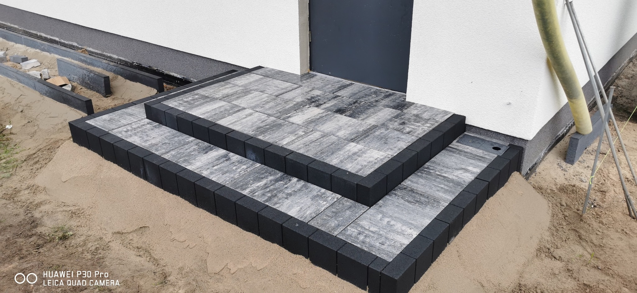 Schody wejściowe do domu wyłożone szarą kostką brukową z czarnym obramowaniem, widoczne ślady piasku po montażu.