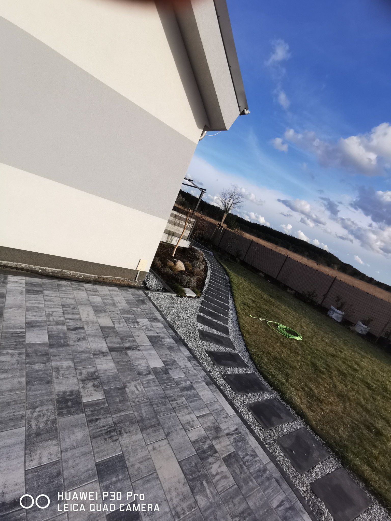 Ścieżka z ciemnych płyt ułożona w trawniku obok domu, oddzielona od kostki brukowej jasnym żwirem, na tle błękitnego nieba z chmurami.