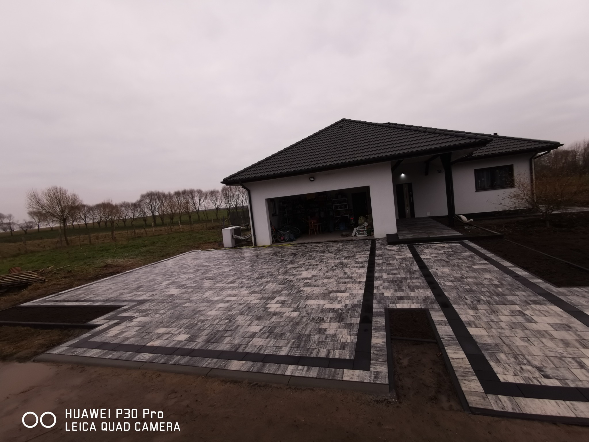 Ułożona szara kostka brukowa z czarnymi obramowaniami przed nowoczesnym domem z garażem, widok z perspektywy ziemi na tle pochmurnego nieba.