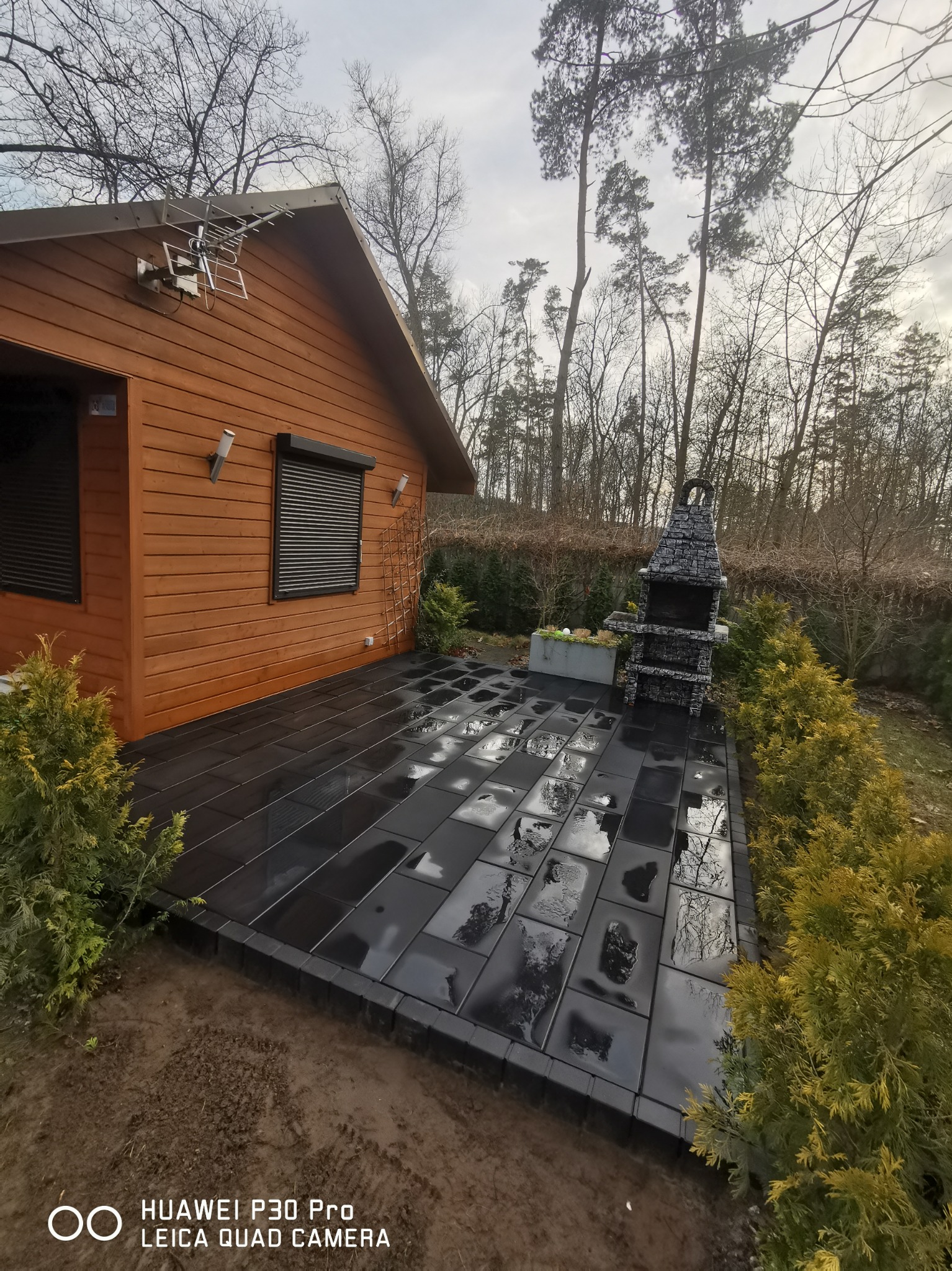 Nowoczesny taras z czarnych płyt, przylegający do drewnianego domku z anteną satelitarną, obok murowany grill i tuje, po deszczu.