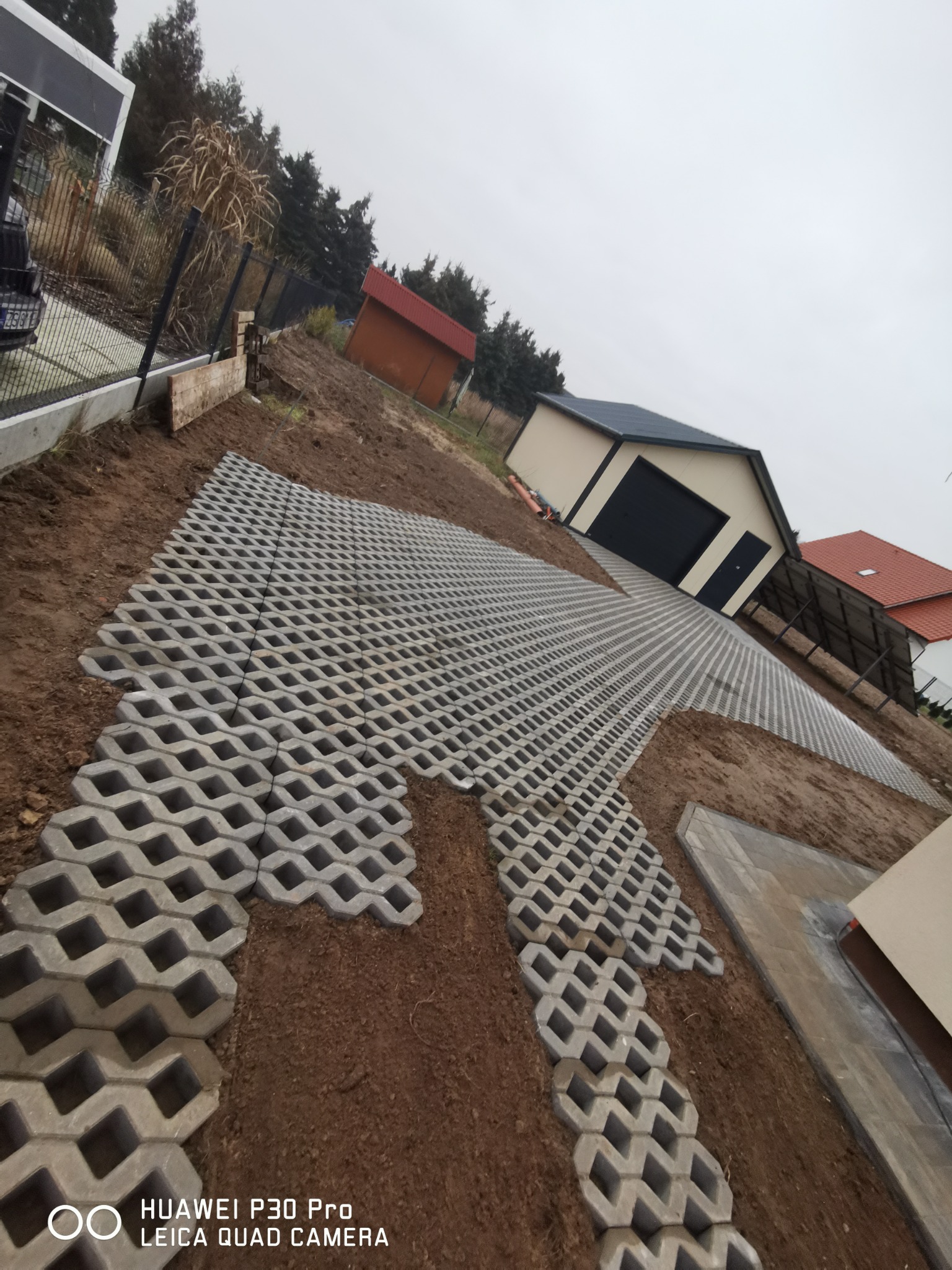 Ułożona częściowo przepuszczalna nawierzchnia z betonowych płyt ażurowych na nierównym terenie działki, widoczny garaż blaszany i budynek gospodarczy w tle.