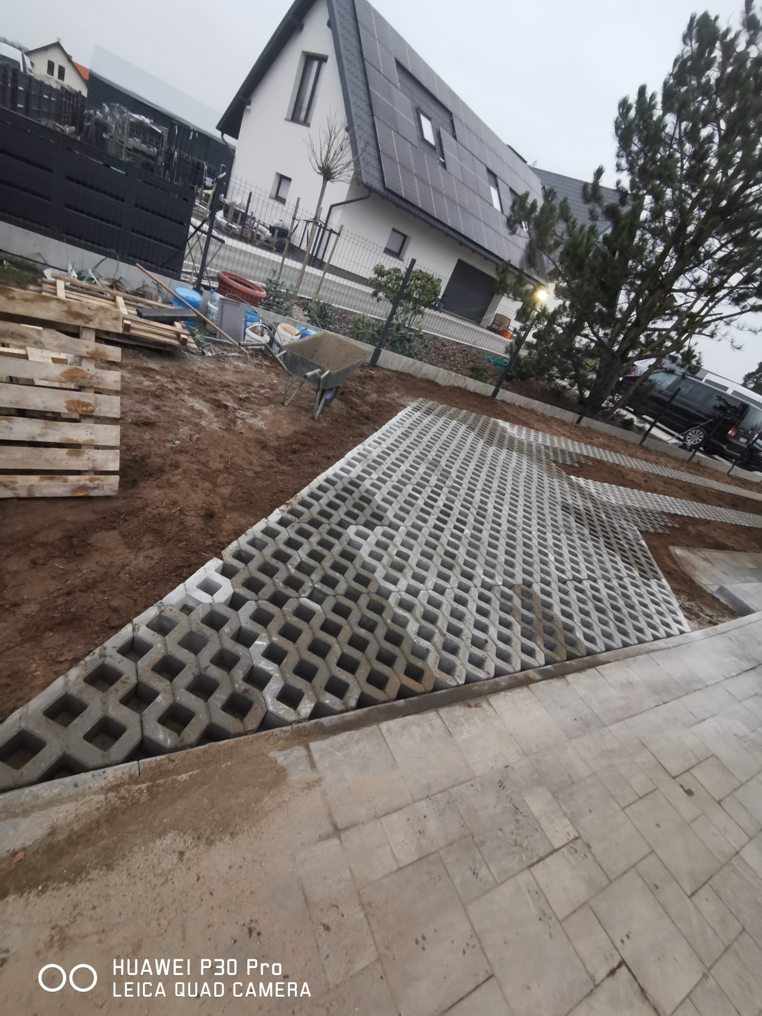 Układanie ażurowych płyt betonowych na podjeździe, widoczne palety z materiałem, taczka i dom z panelami słonecznymi w tle.